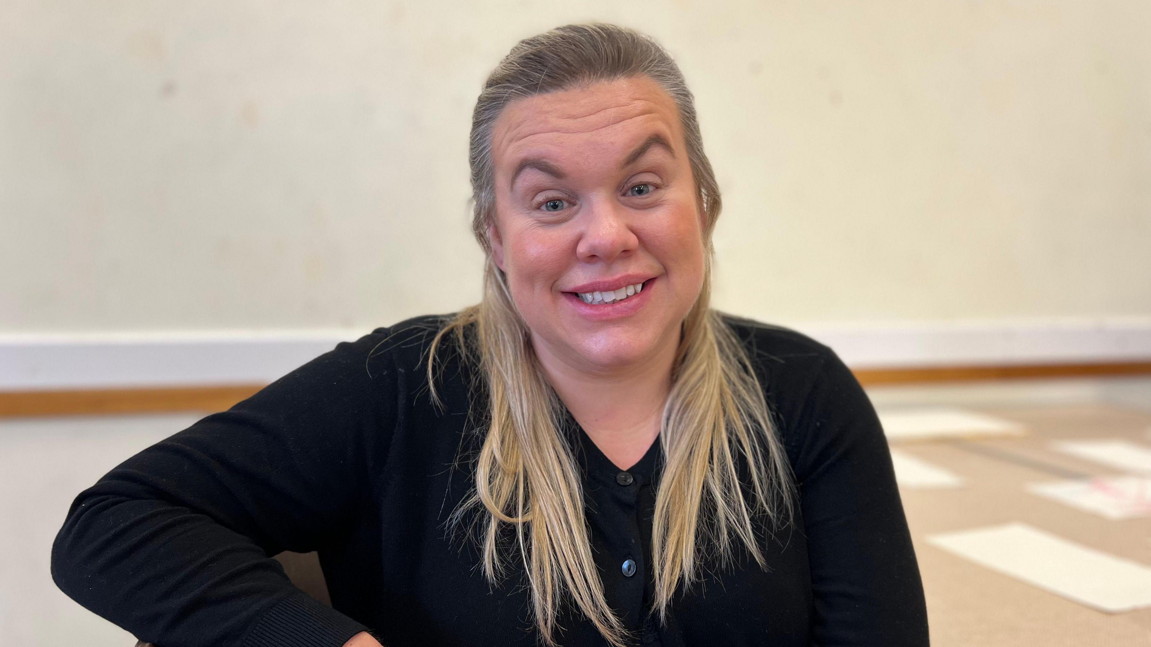 Amber Regnard pictured with long straight blonde hair. She is smiling at the camera and wearing a black cardigan. 