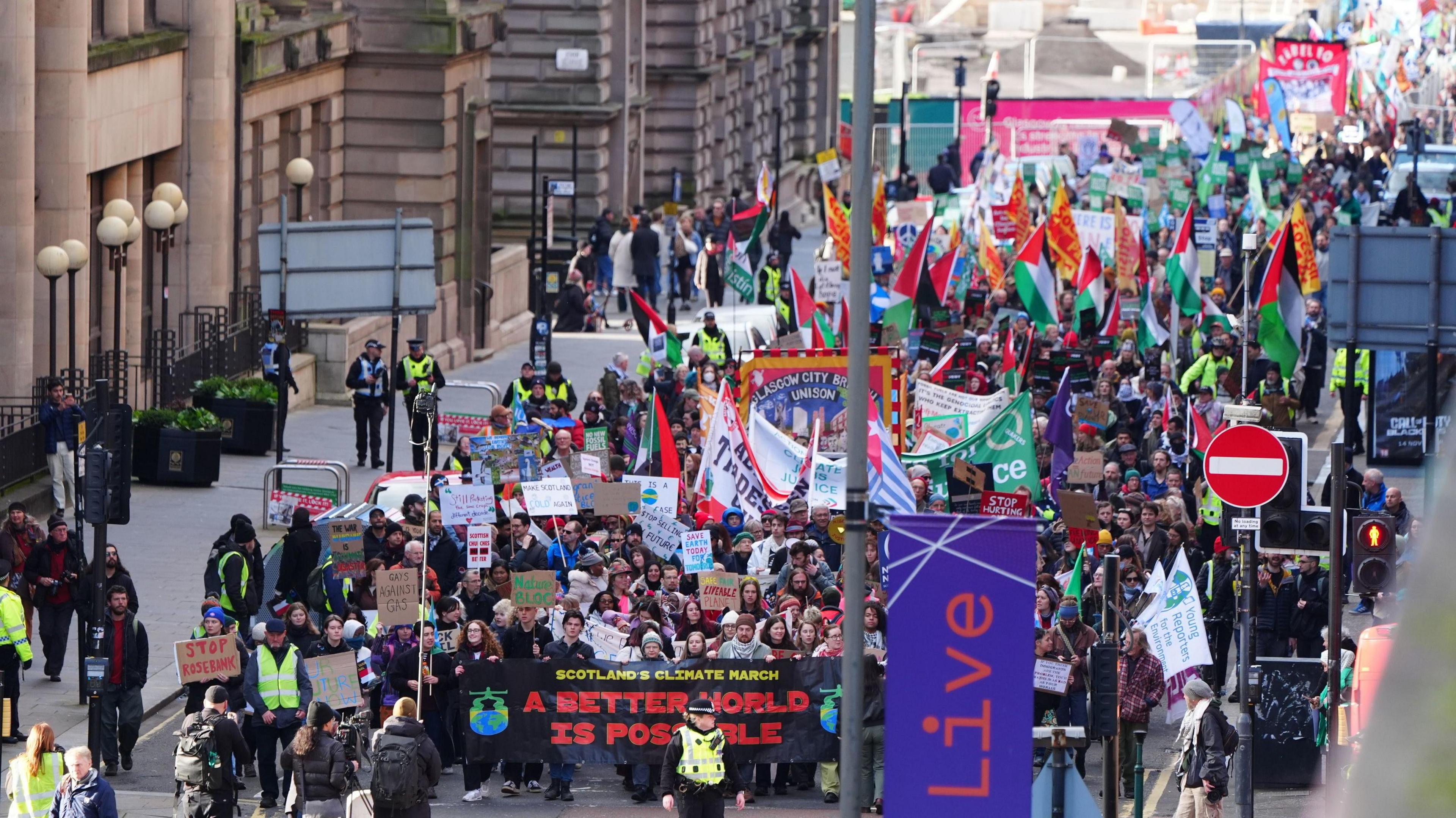 A wide overhead view of a large climate march filling a city street in Glasgow. Hundreds of people carry banners, placards and flags, including a large front banner reading “A Better World Is Possible.” Many signs reference climate justice, Gaza, and environmental protection. Police officers line the route, and tall buildings rise on both sides of the street as the crowd stretches far into the distance.