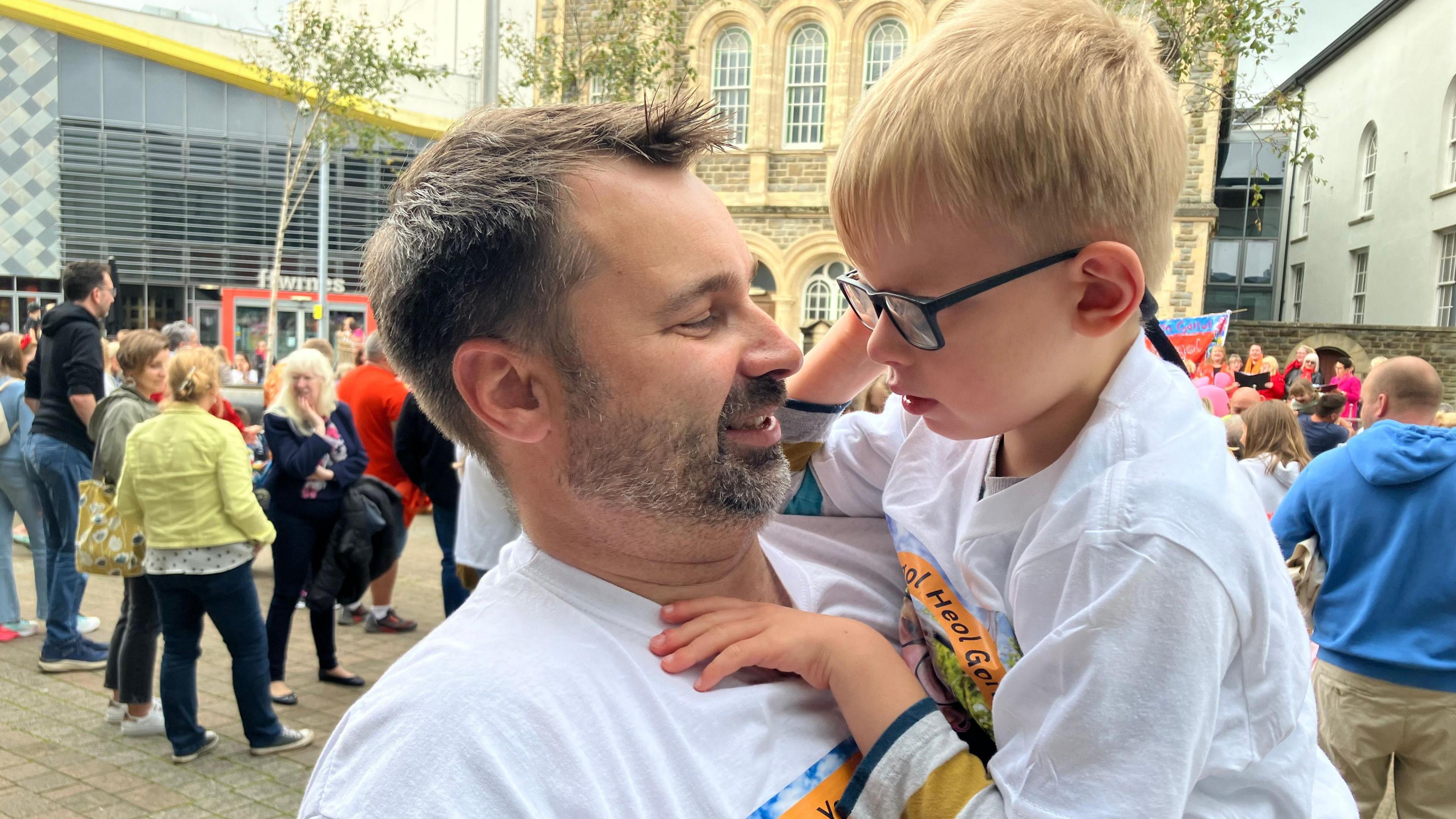 Alex holds his son on his hip. They are look at each other. Alex has short dark hair and a dark beard with grey. His son is blonde with dark-rimmed glasses. Both are wearing white t-shirts.