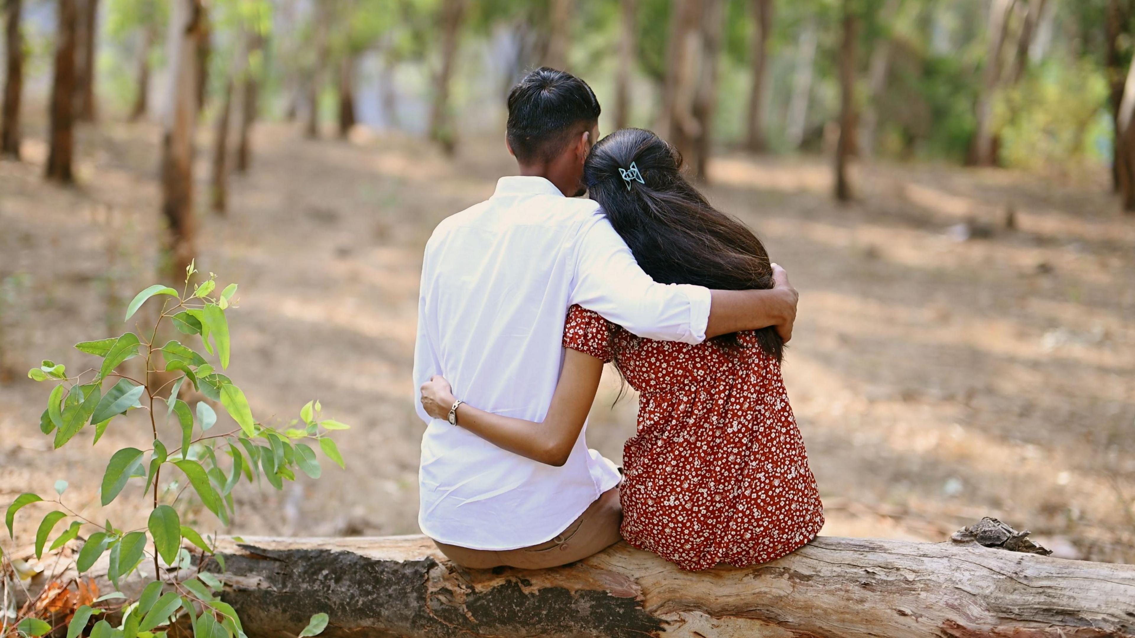 A boy and girl put their arms around each other in a garden in India. The boy is wearing a white shirt while the girl is wearing a maroon tunic with pattern. 