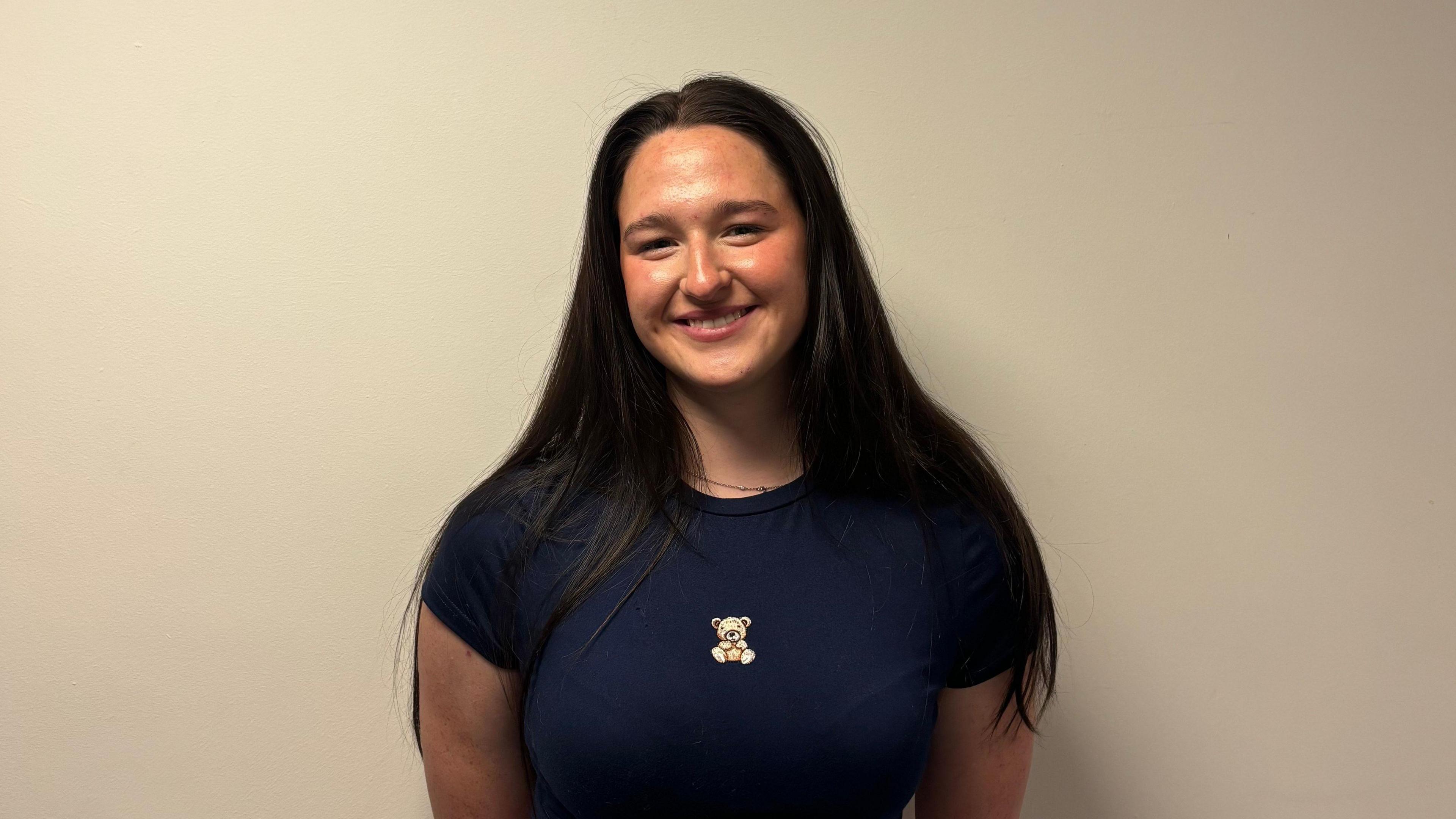 jessika is standing in front of a white wall. She is wearing a navy top with a teddy bear on it. She has long black hair. She also has a small chain around her neck.