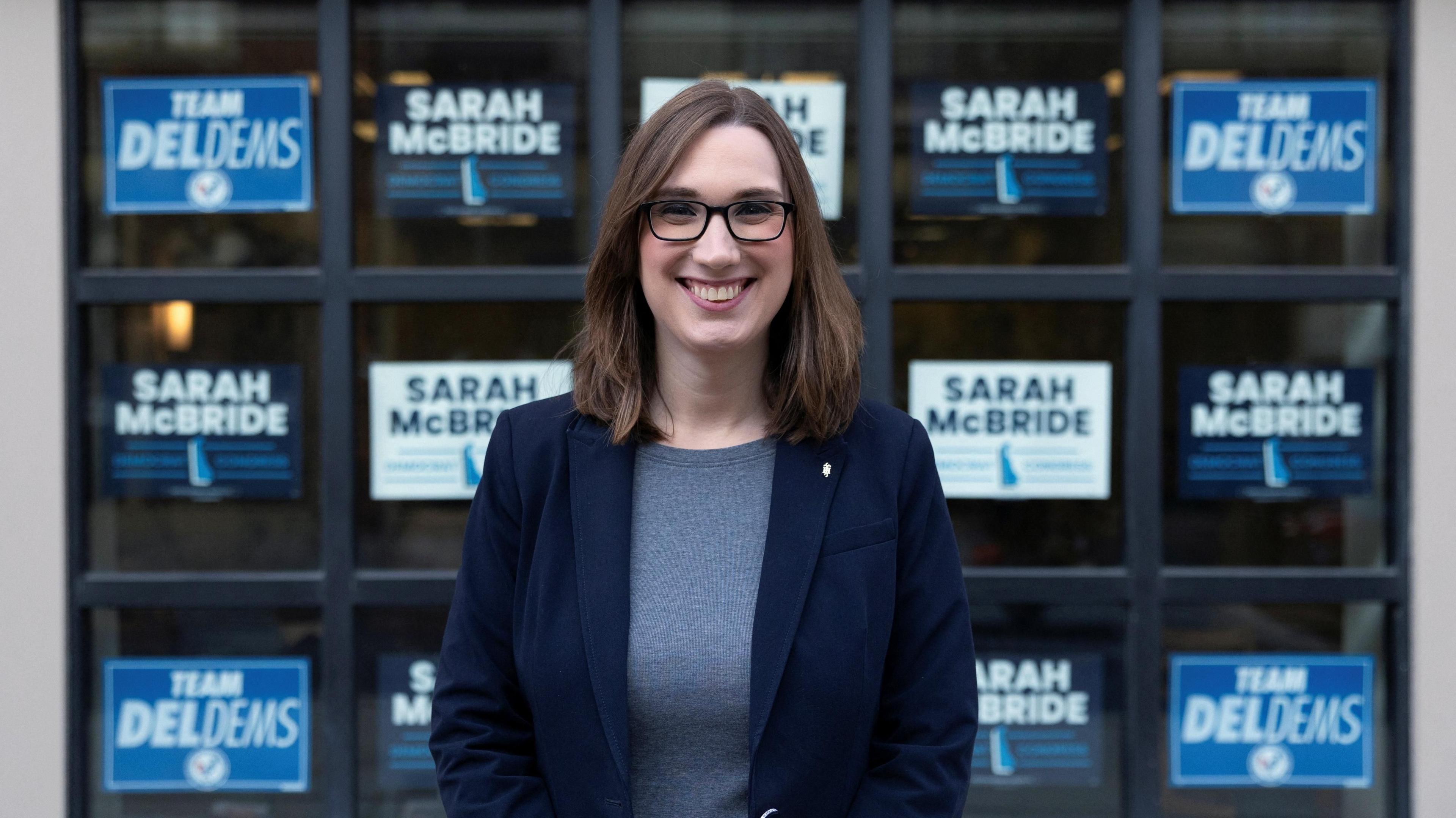 Sarah McBride, Democratic member of the senate for Delaware, stands in front of signs saying her name and DelDems. She wears a blue blazer and grey top