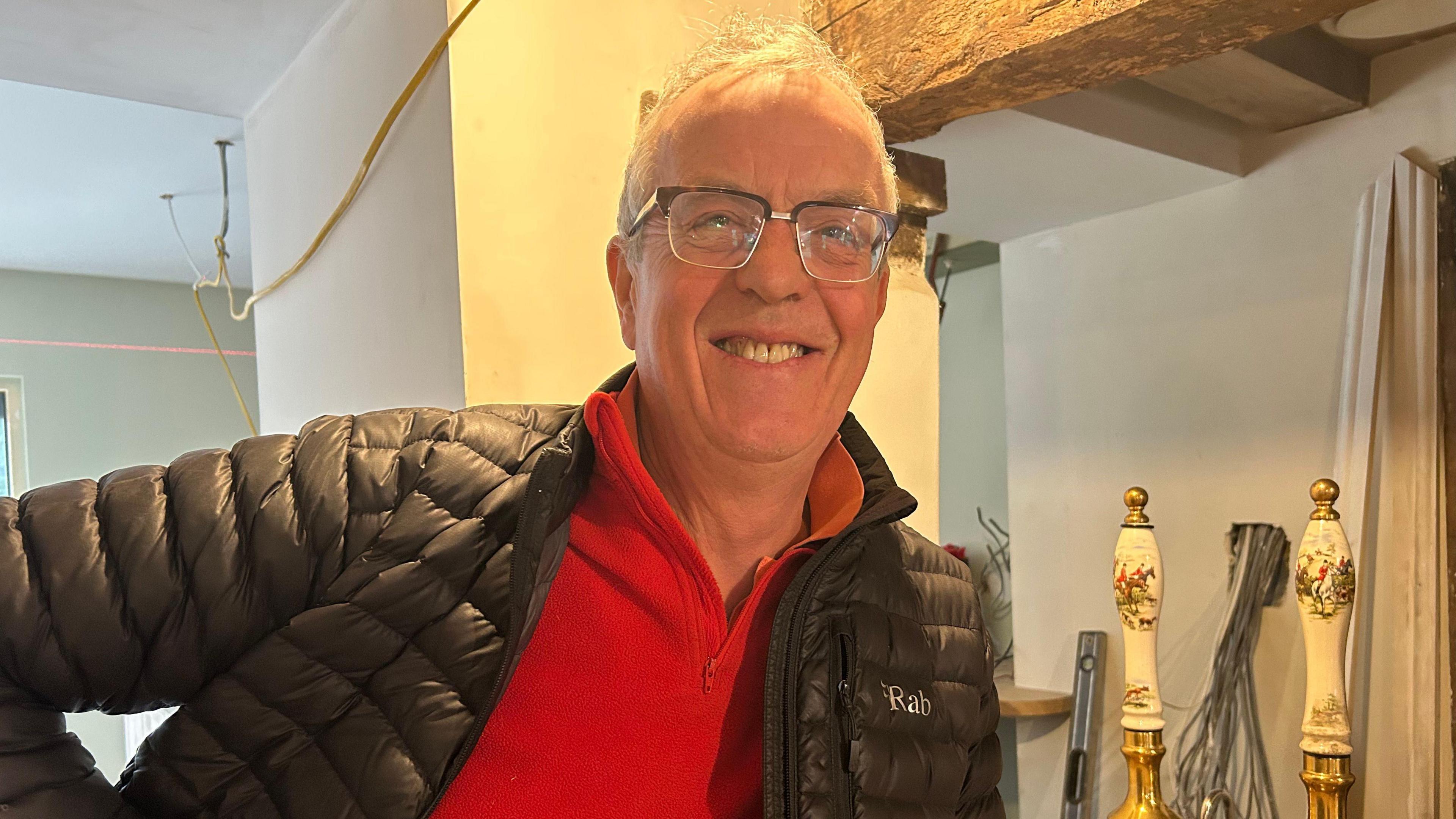 Simon Coombe wearing glasses, a red three-quarter zip jumper and black padded jacket. He is leaning on the newly installed bar at the Hop Pole Inn. Construction is still under way, and in the background there are white mouldings leaning up against the wall and wires hanging dangling from the ceiling.