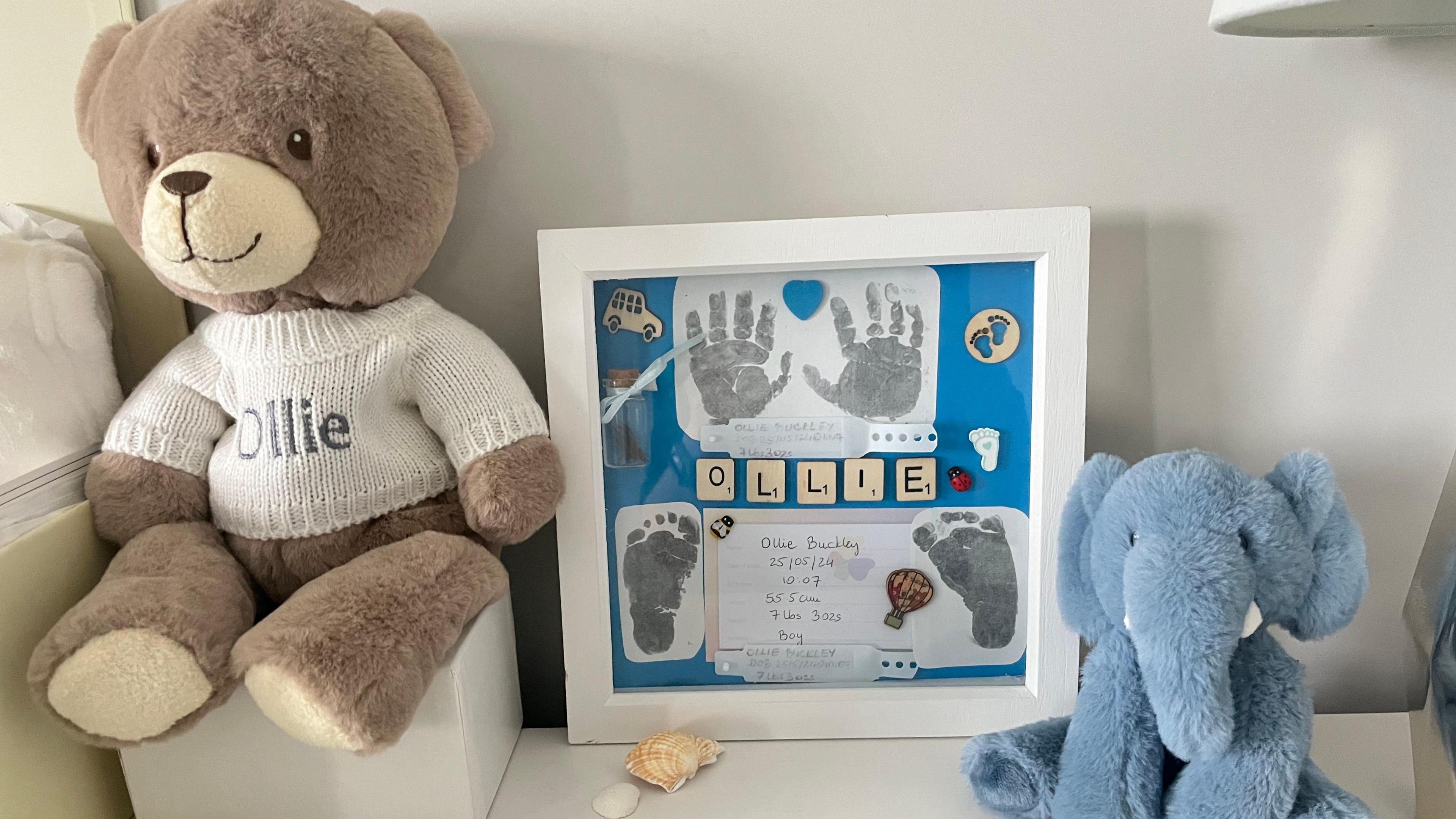 A brown bear with a jumper saying "Ollie" sits beside a frame which has painted grey hand and footprints of baby Ollie. A blue elephant plush toy is beside the frame along with two shells from the beach.