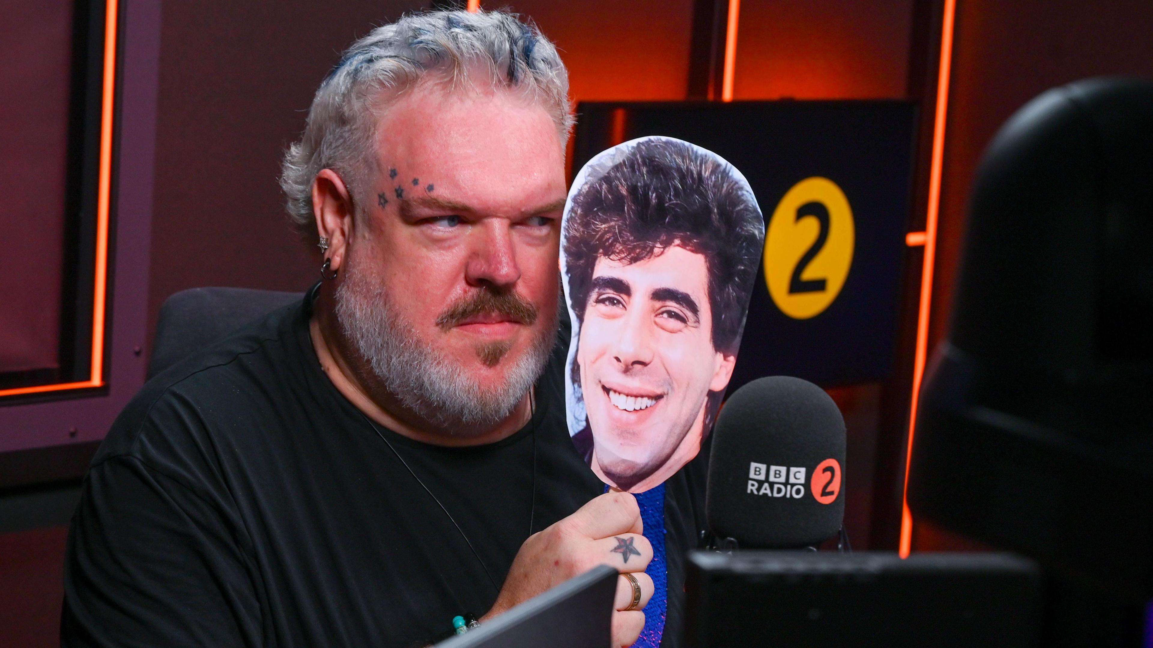Kristian Nairn looking out from behing a mask of Gary Davies's face in the radio studio with Radio 2 logos