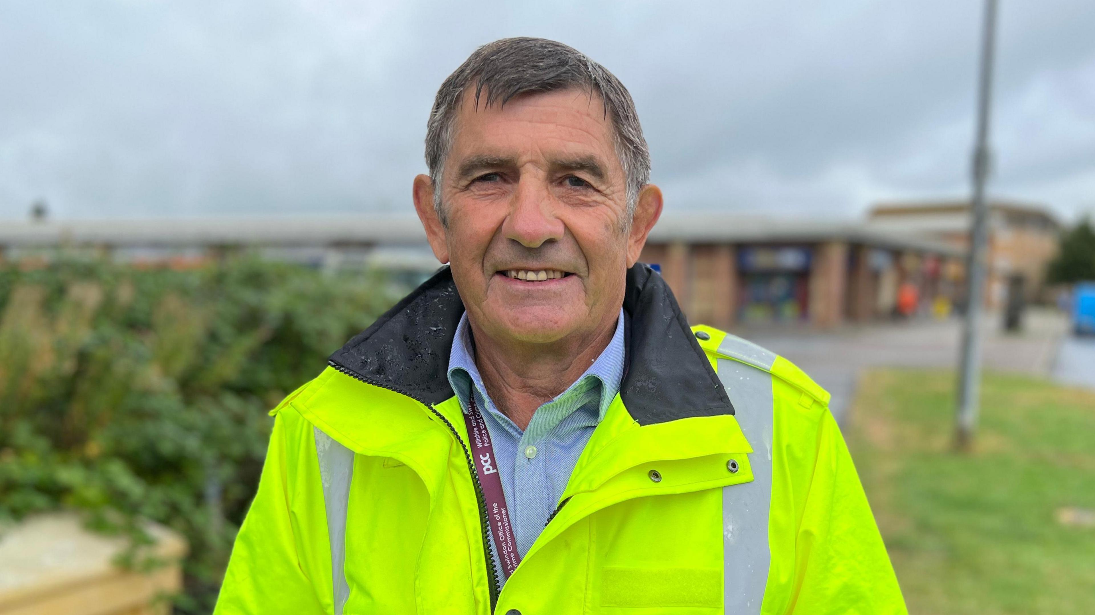 Mr Wilkinson, shown wearing a yellow high-vis jacket, a blue shirt and a purple lanyard with 'PCC' written on it. He has short grey hair and is shown smiling at the camera. 