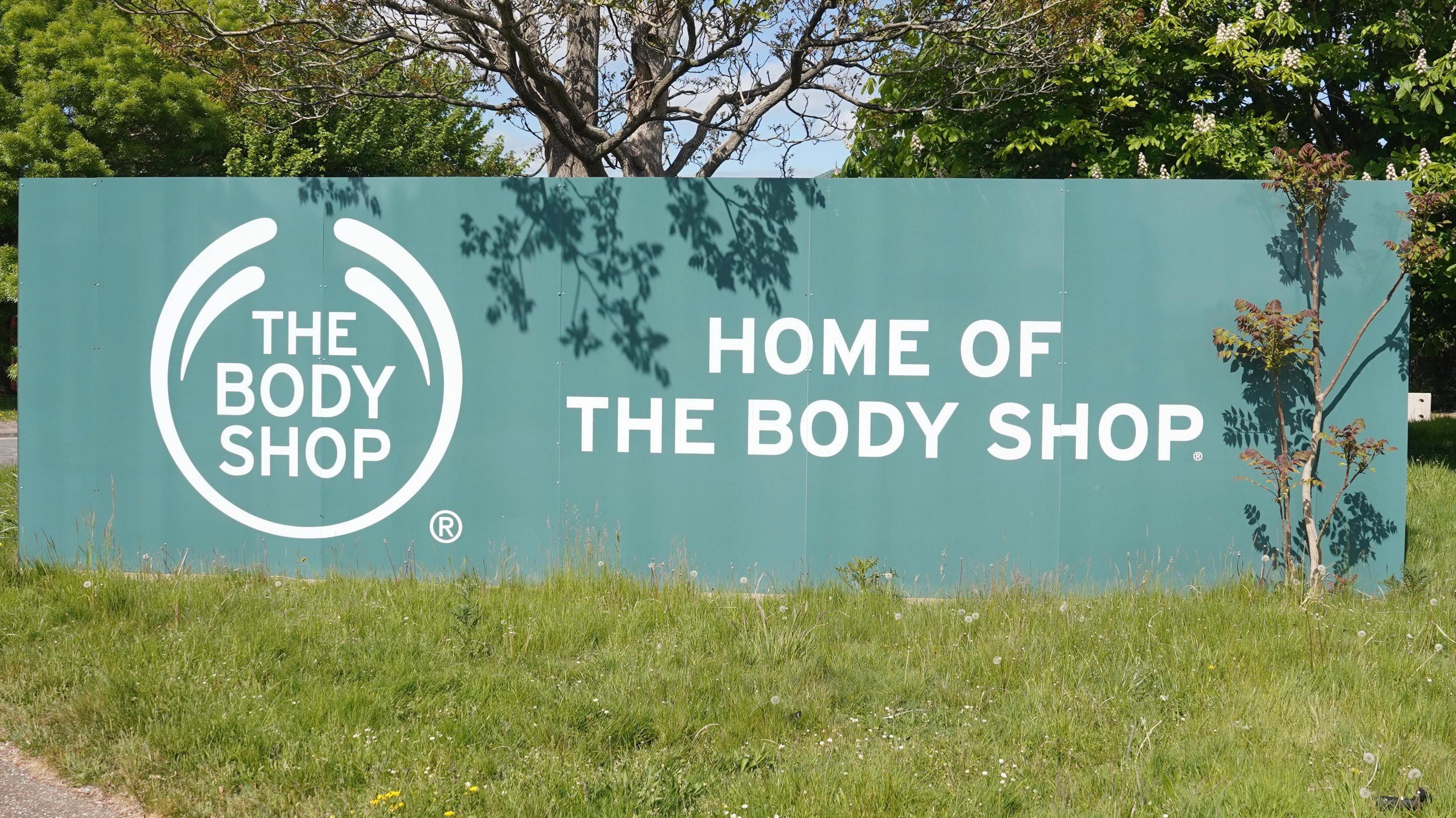 A dark green sign with writing and a logo on reading "Home of The Body Shop".