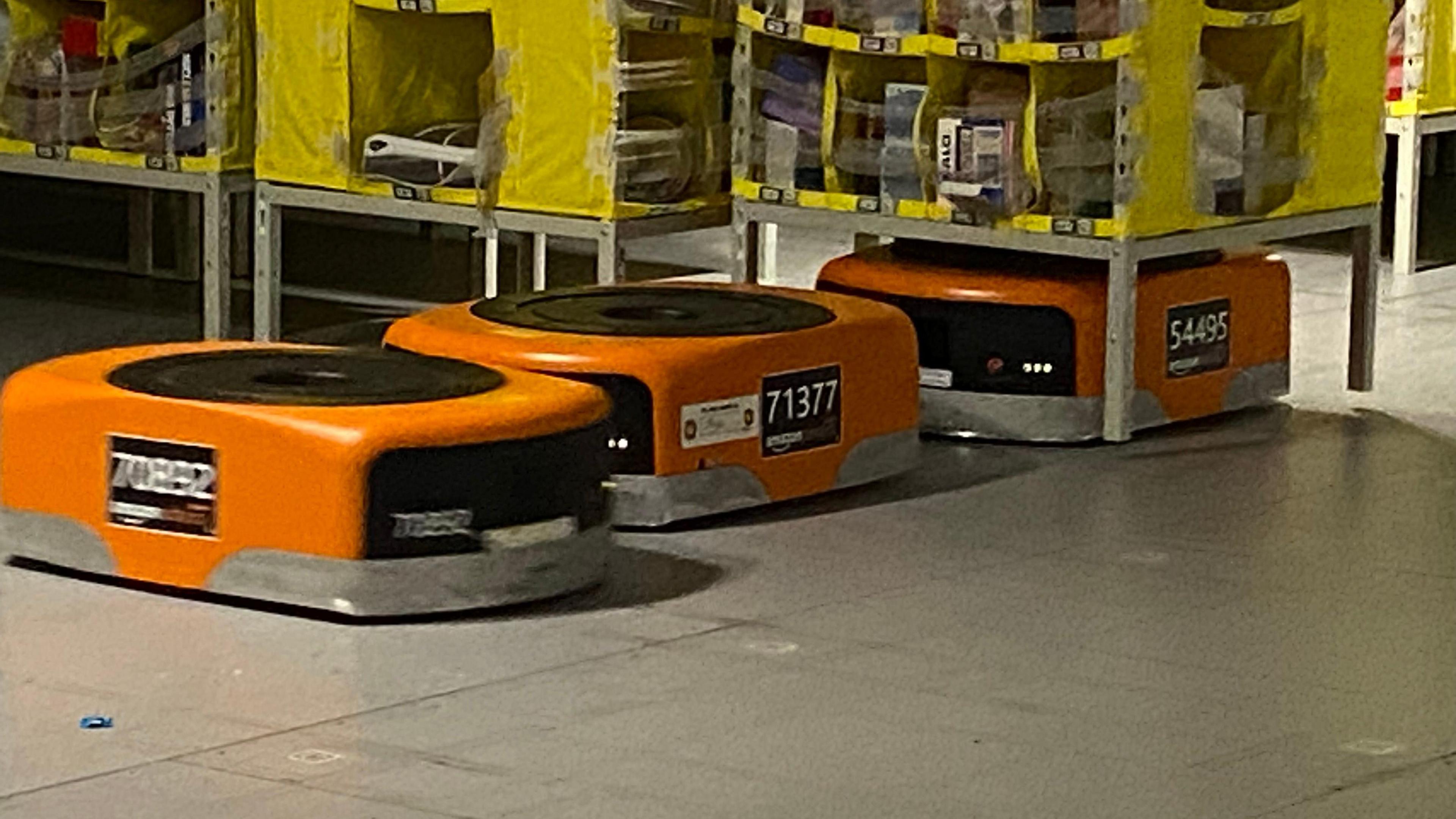 Orange robots, the size of small boxes, run along a warehouse floor, under yellow shelving stacks, which they run under, attached to, and move around. 