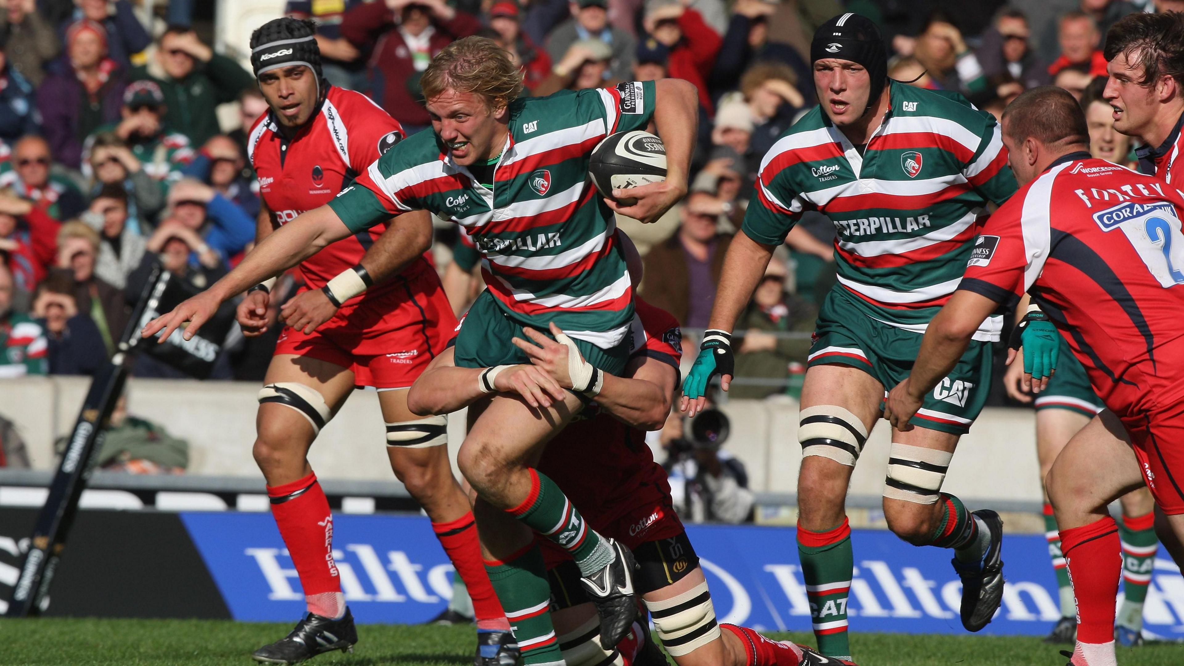 Moody is being tackled on a rugby pitch while being surrounded by other players in red shirts. He is holding the ball and is in a green, red, and white shirt.