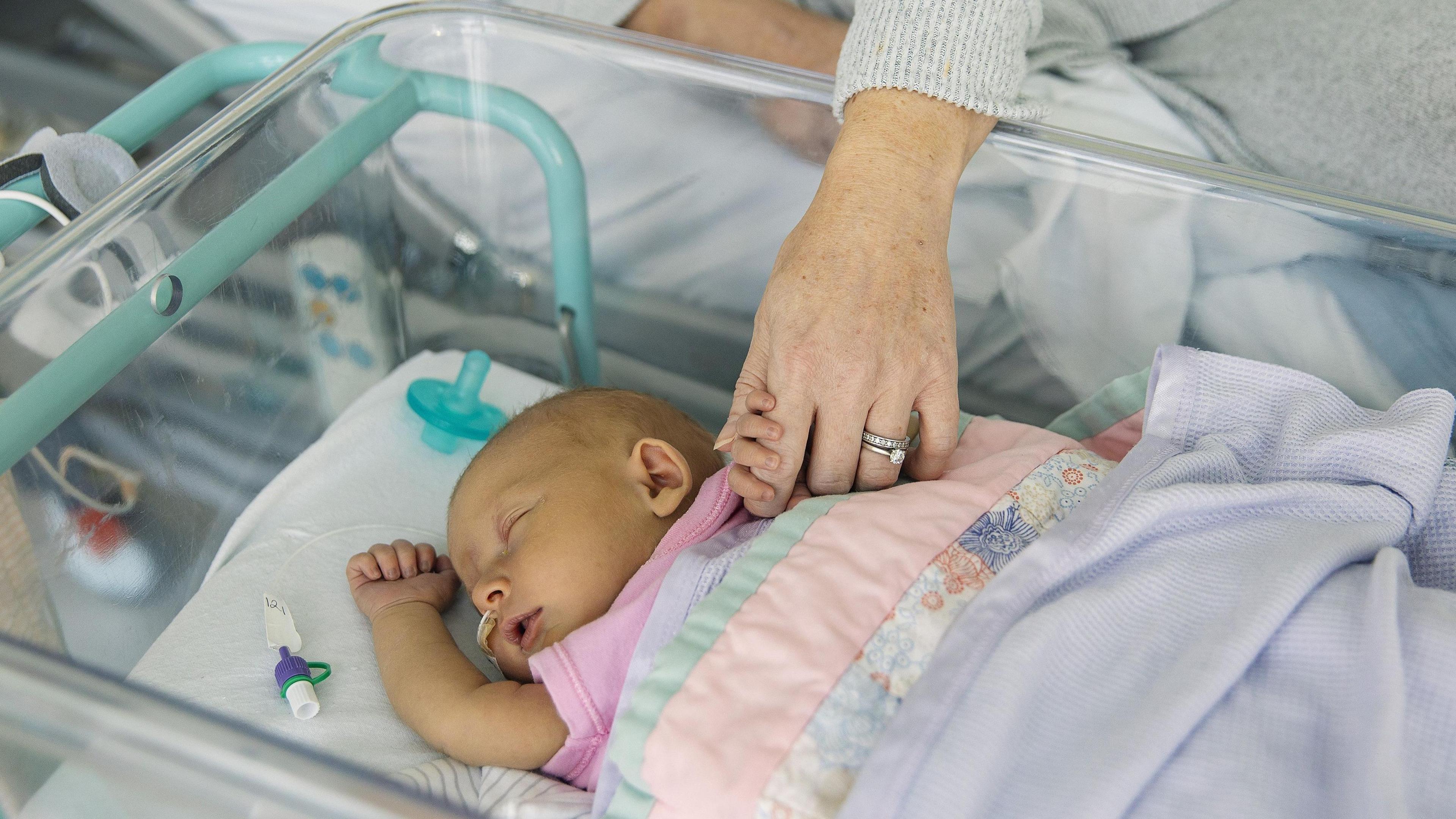 A newborn baby sleeps in a bassinet while its mother, lying in a hospital bed, holds one of its hands.