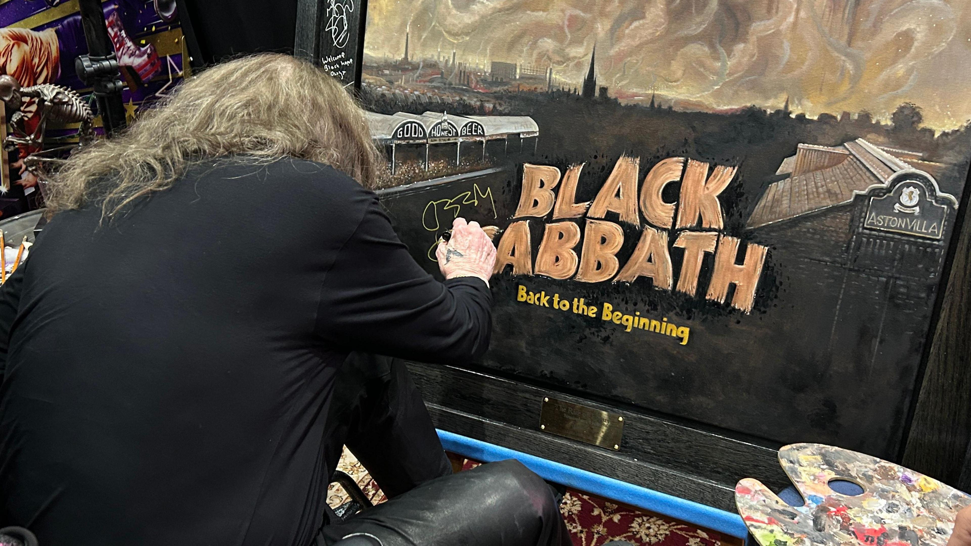 Ozzy Osbourne is leaning forward to sign his name on the Black Sabbath painting.