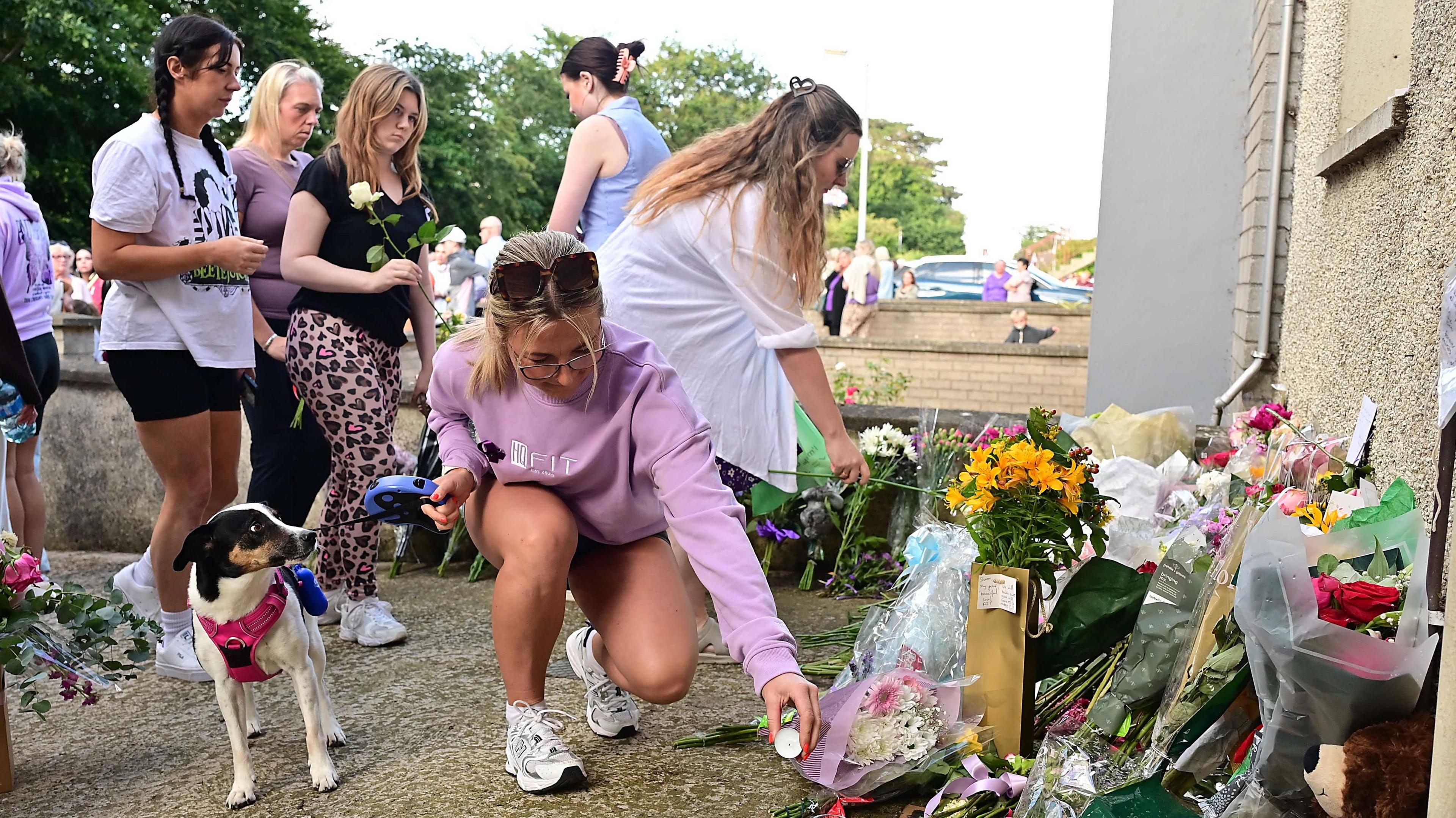 A woman placing a candle on the ground beside a pile of flowers. There are five women behind her and a dog beside her. 