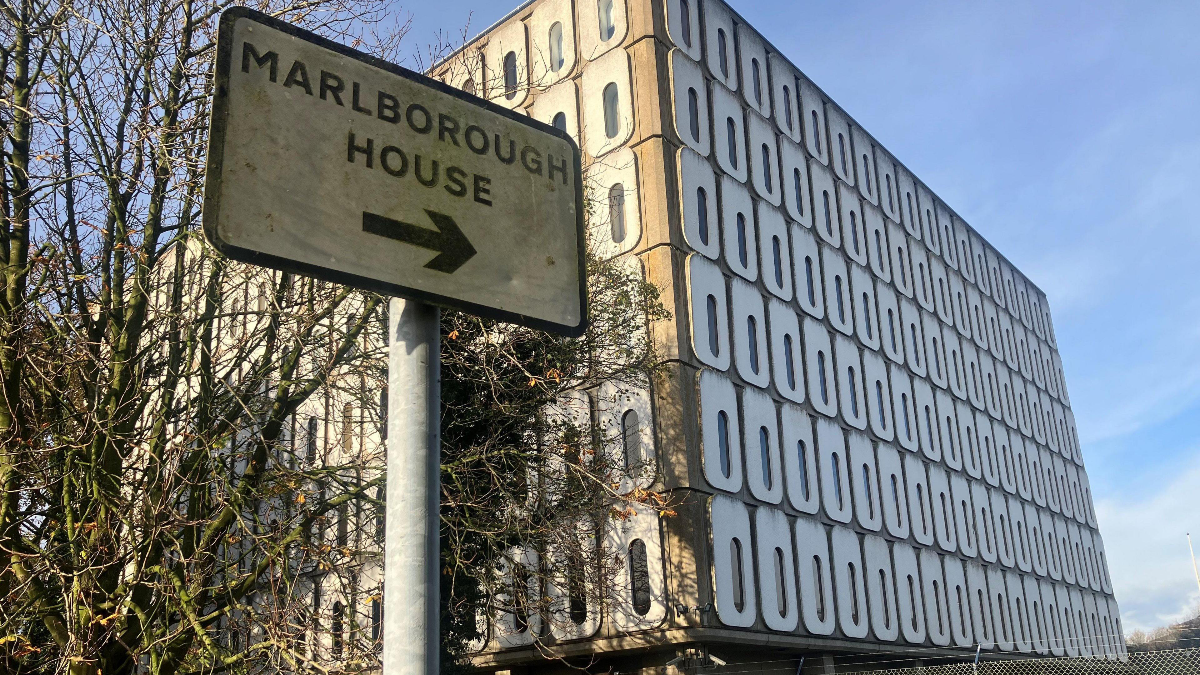 A sign saying Marlborough  House in front of a square grey building with rows of thin oval shaped windows.