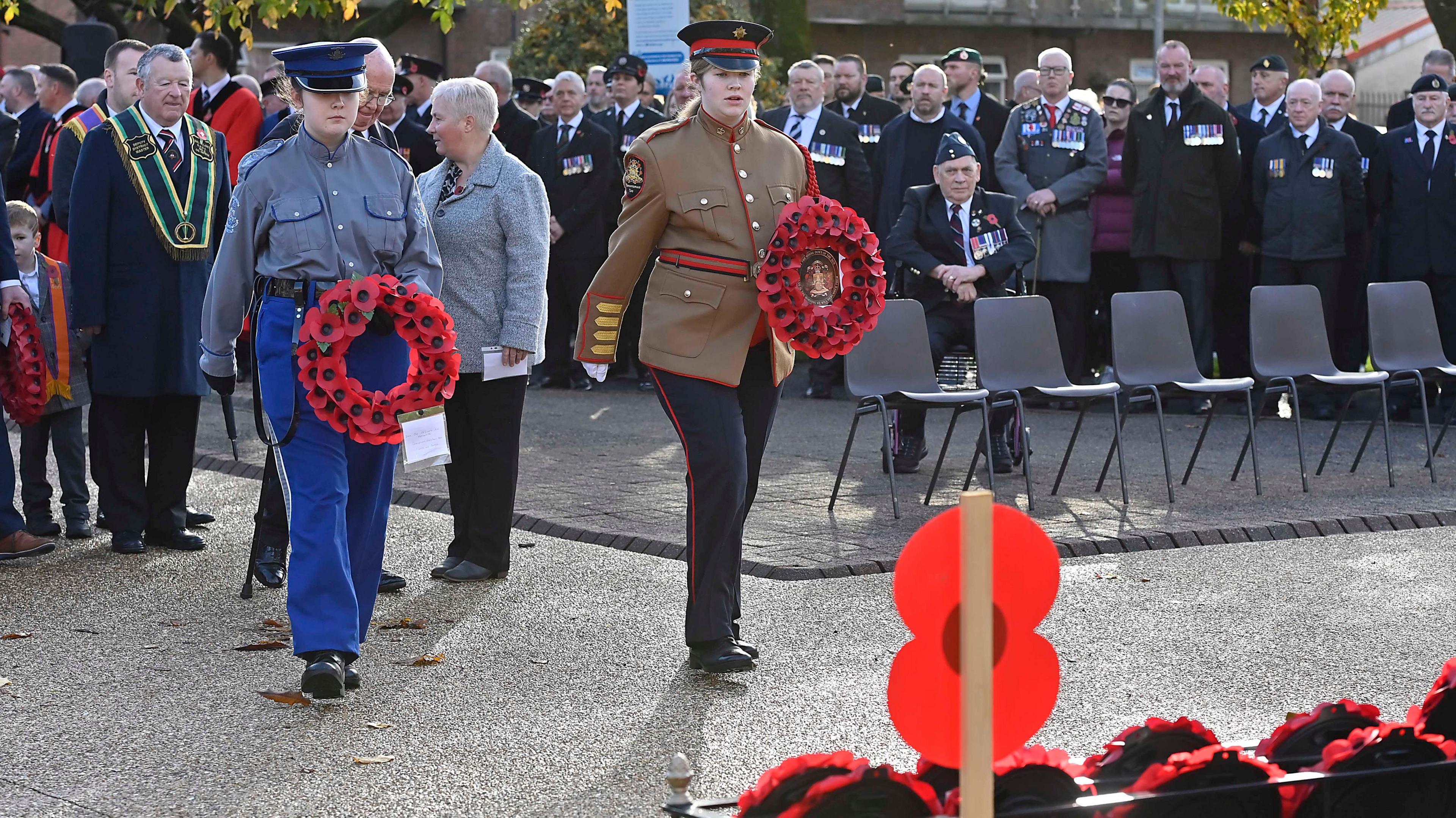 Young people wearing uniforms are holding poppy wreaths. There's a crowd behind them. In front of them of a memorial with wreaths. 