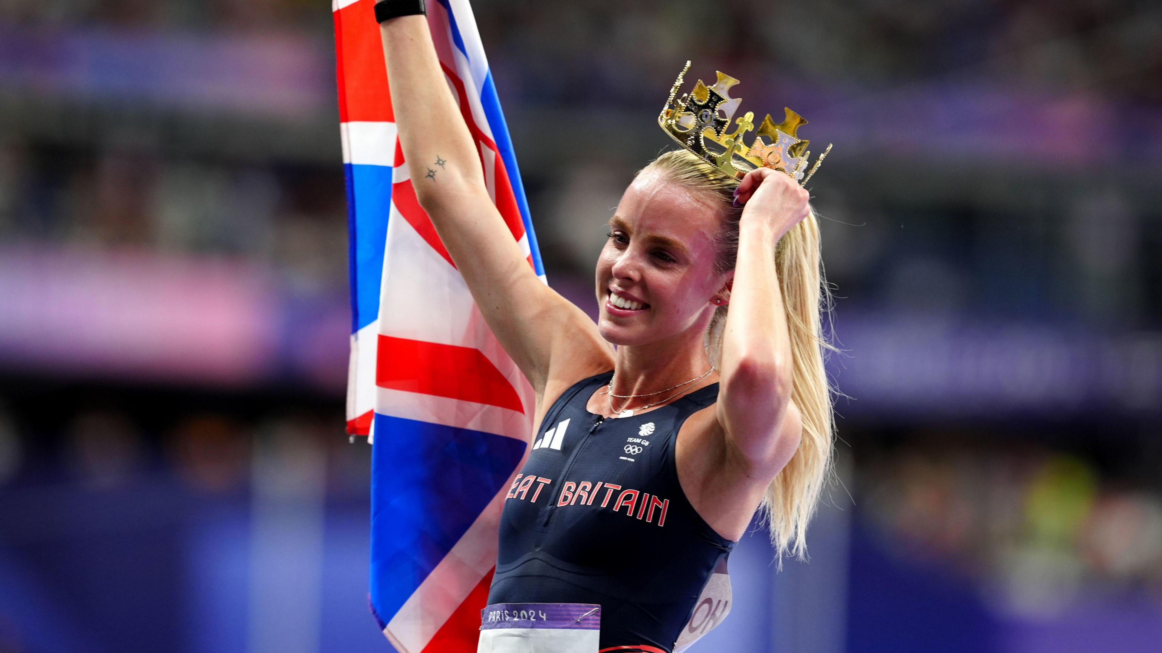 Keely holds up a union jack flag and with the other hand, holds a plastic gold crown to her head. She is wearnig a Great Britain jersey