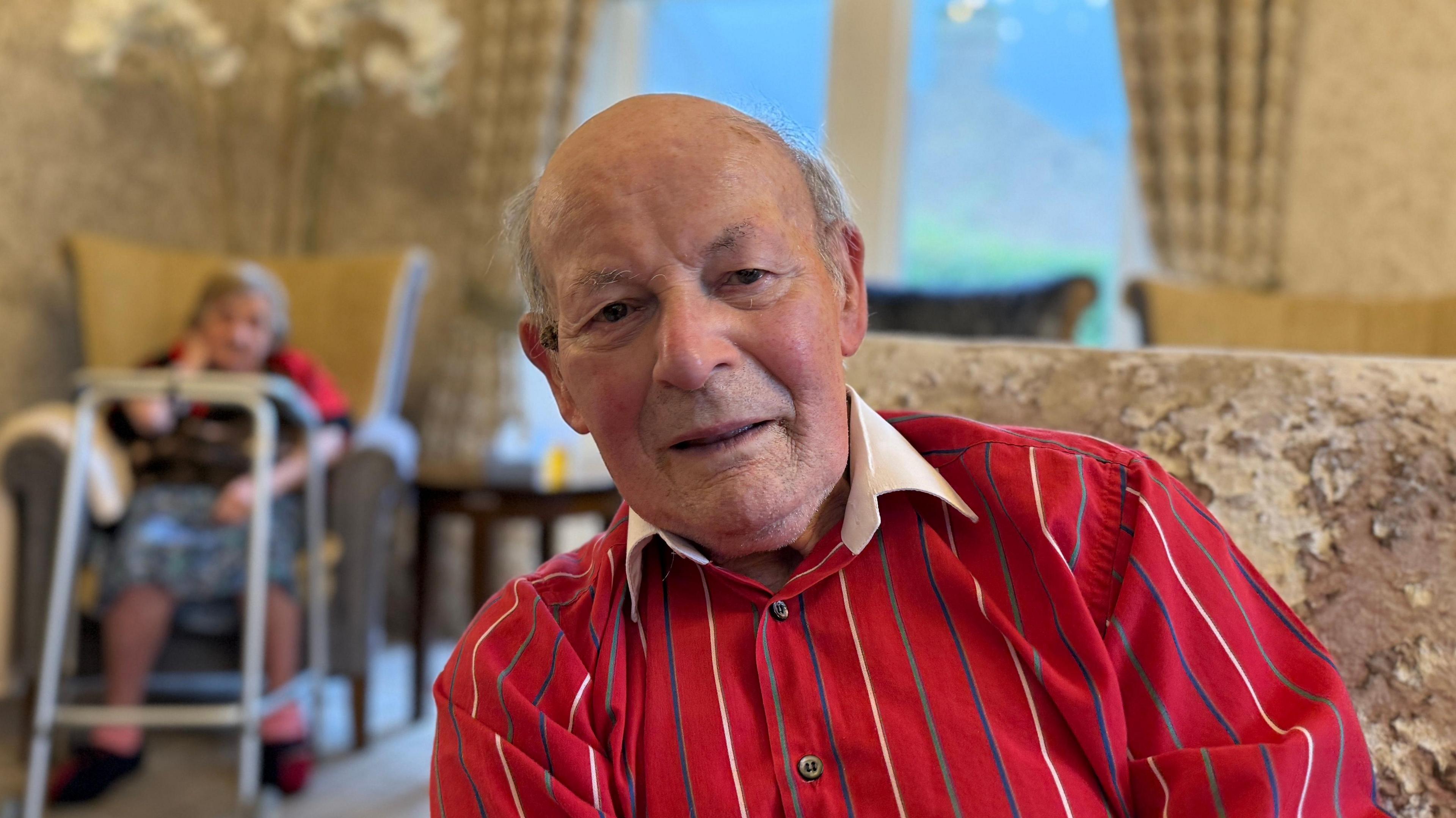 A man sat in a chair wearing a red shirt with white, green and blue thin vertical stripes on it and a white collar. Top button undone. Blurred background shows a beige room. 