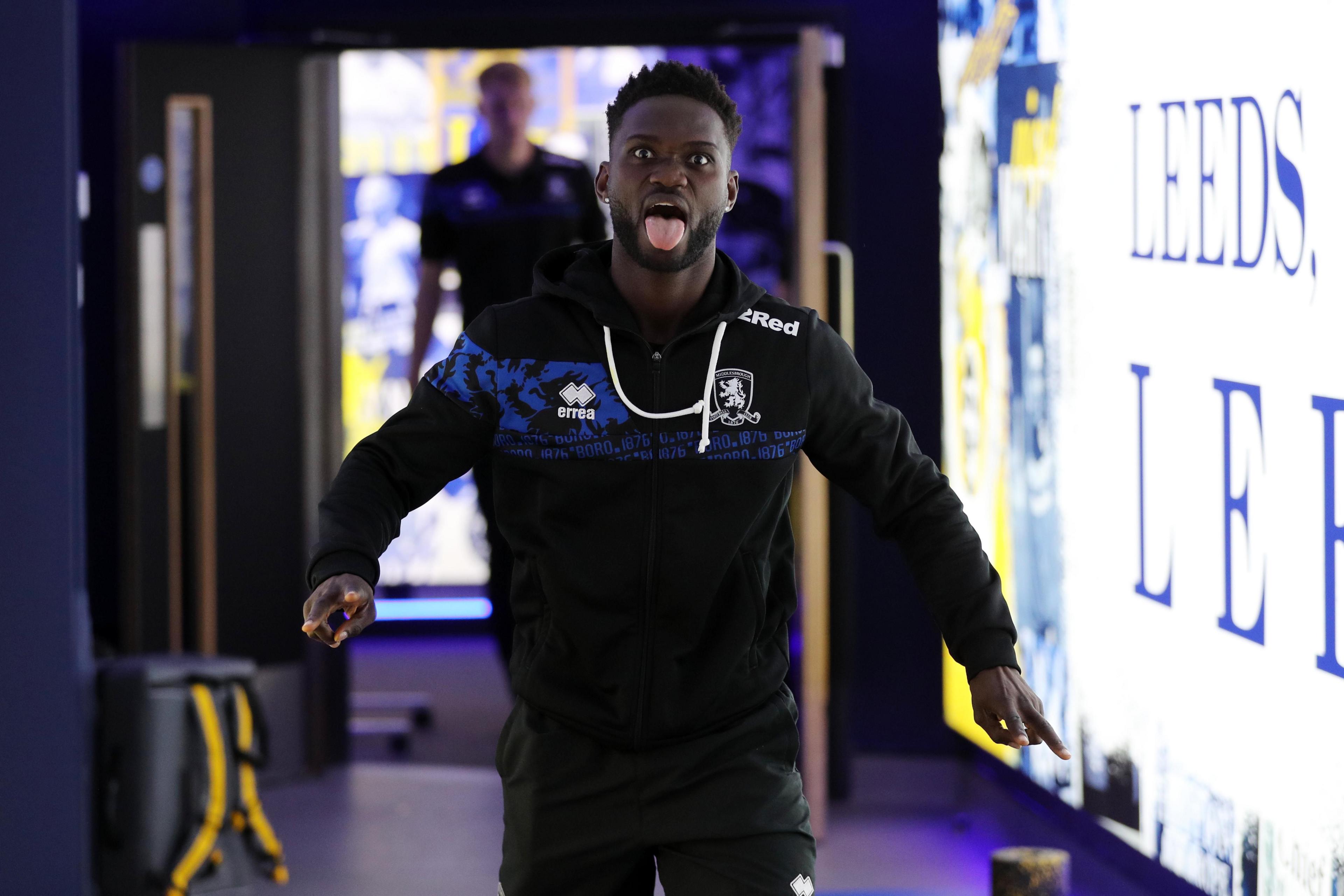 A footballer in a black tracksuit sticks his tongue out as he walks towards the camera next to a large illuminated sign that reads "Leeds".