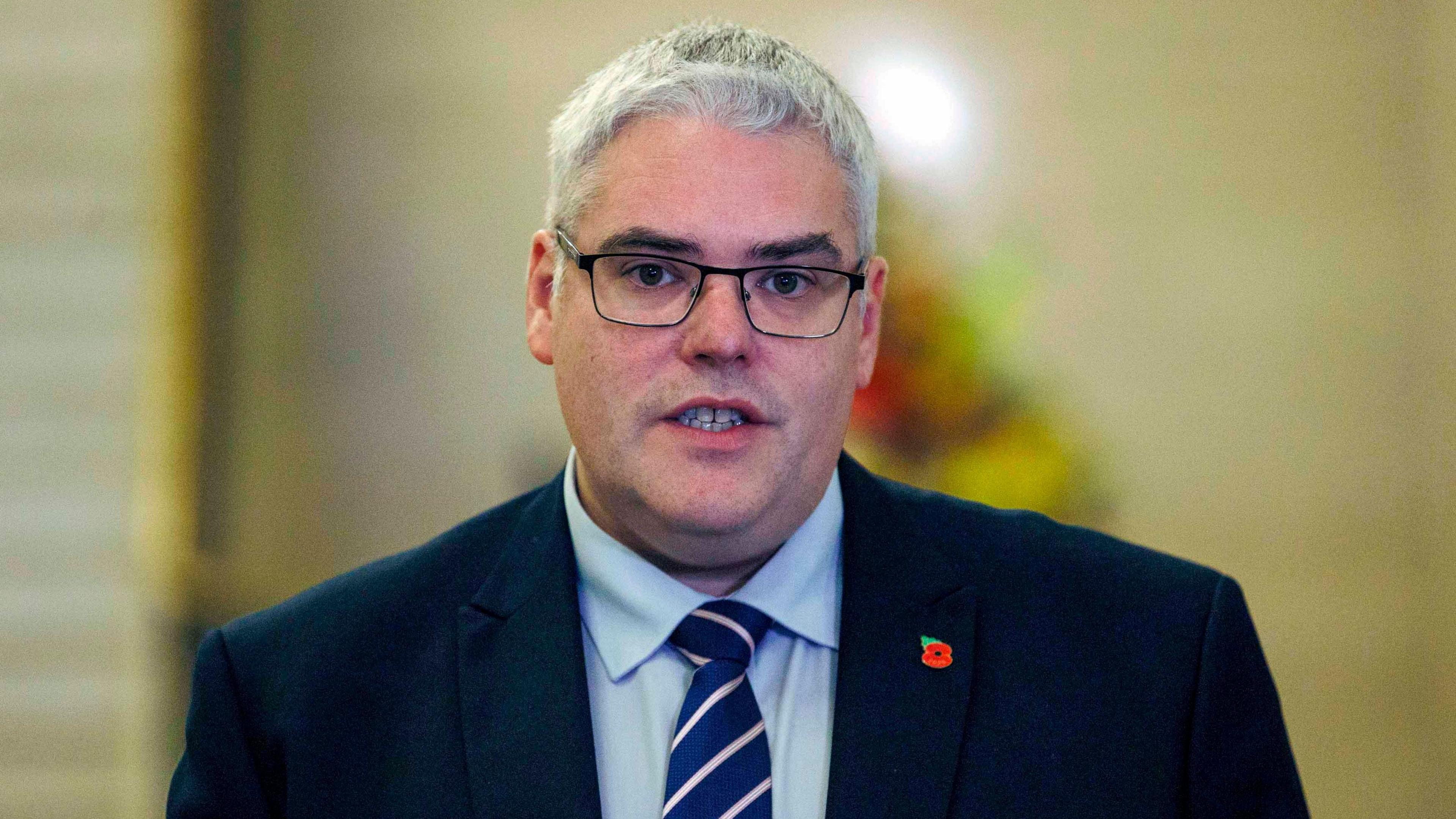 DUP leader Gavin Robinson speaks to the media during a press conference in the Great Hall, Parliament Buildings, Stormont. He is wearing glasses, has on a blue and white tie, blue shirt and black suit coat with a remembrance poppy pinned. 