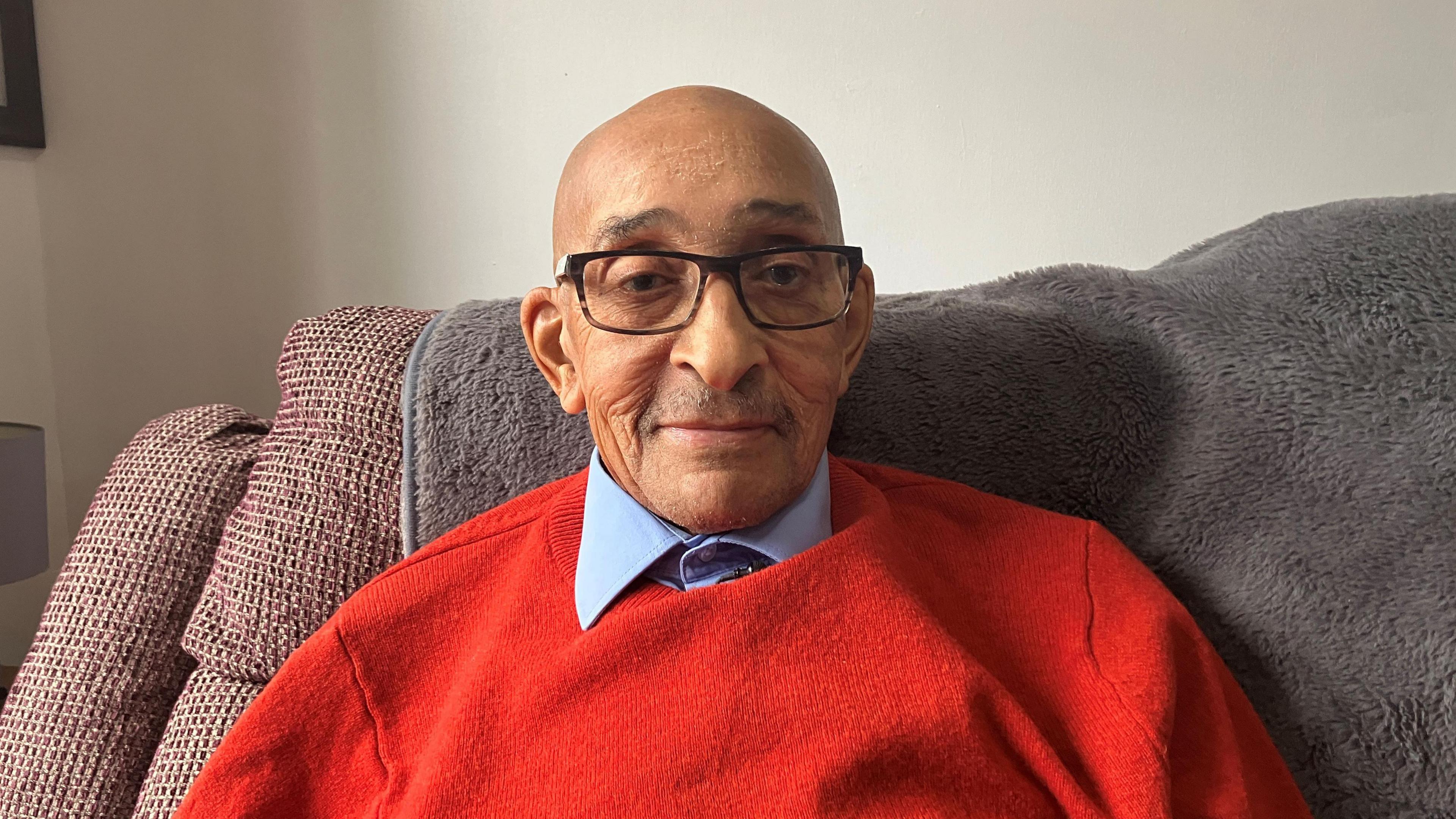 A man in a red jumper and blue shirt wearing glasses and with a bald head. He is sat on a sofa chair which has a grey furry throw on it.