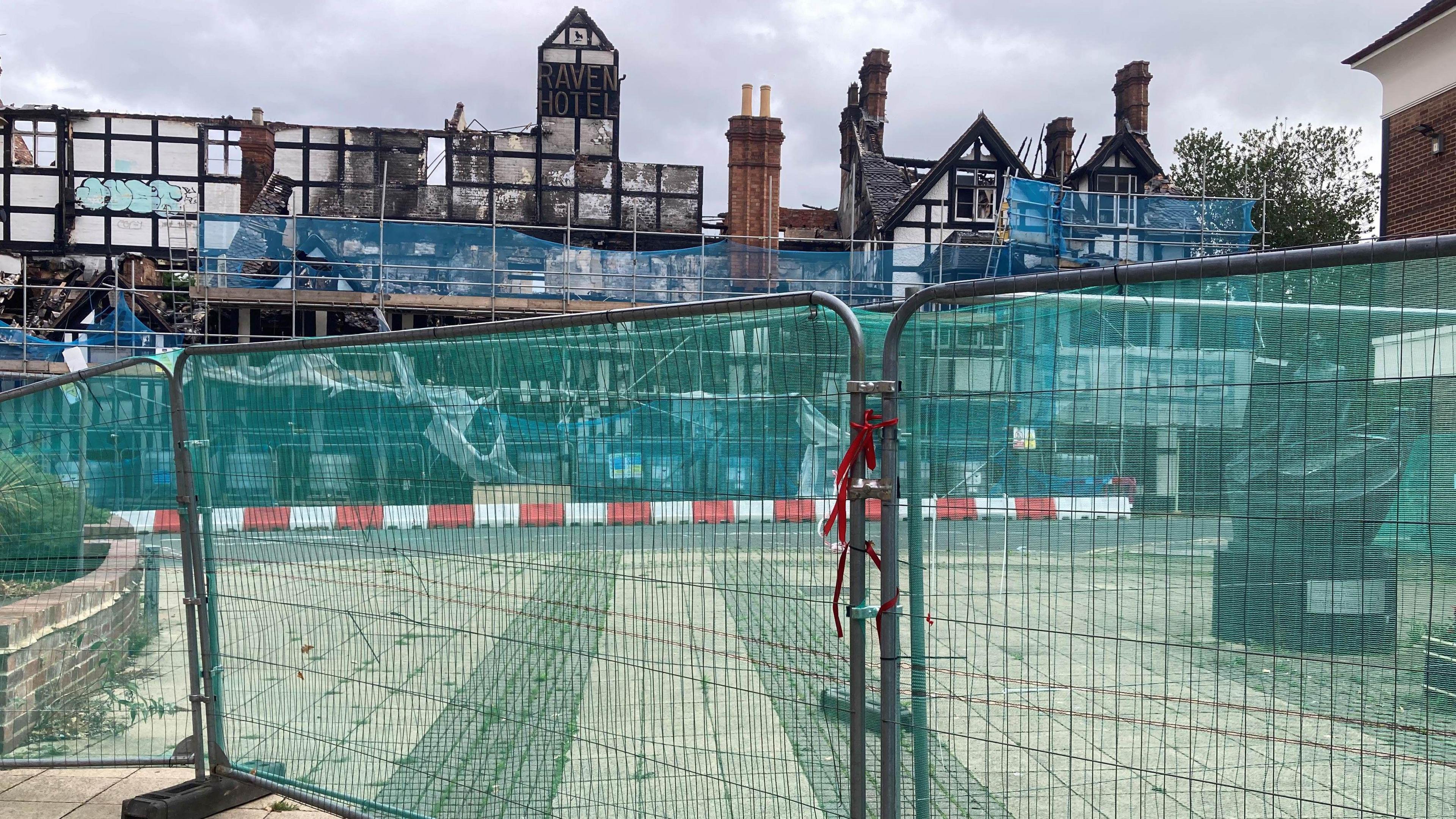 A fence in front of a large black and white building, which has smashed windows and visible blackened-out sections after a fire. 