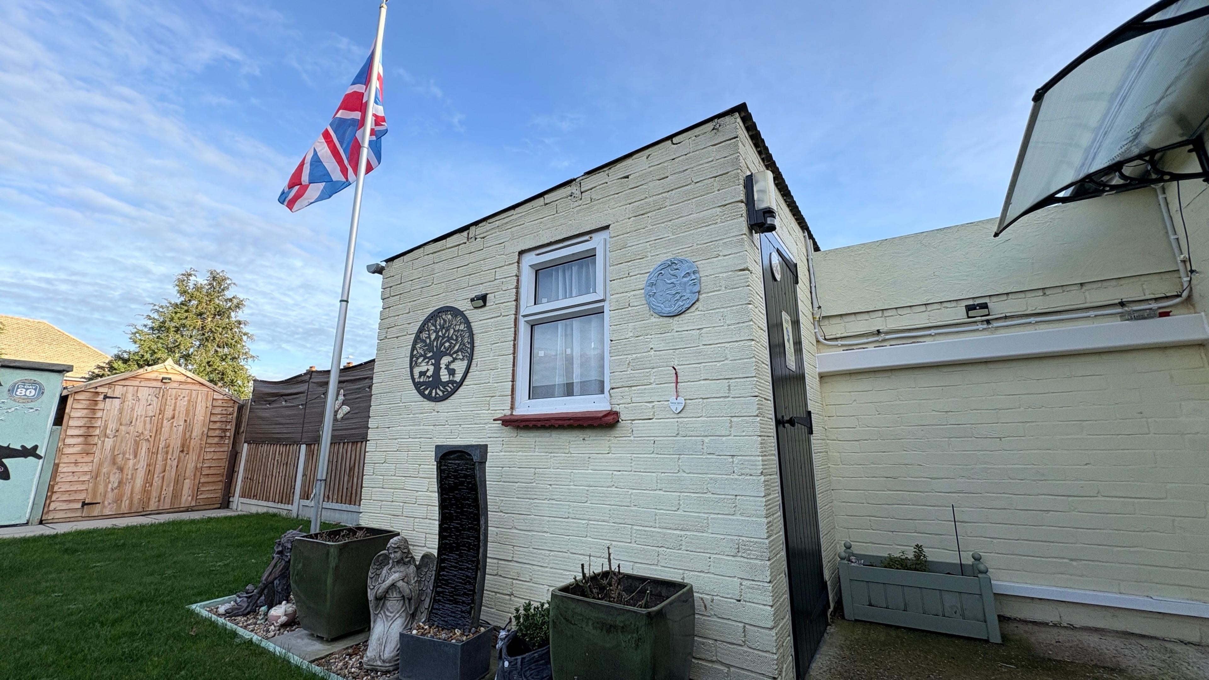 A brick built shed, with cream walls, in a garden. We are looking at the side wall of the shed, which has a small window in it, through which we can see a net curtain. A black door into the shed is just visible in the front wall to the right. There is an area of grass to the left of the image and behind the shed. A flag pole also stands behind the shed, from which the Union Flag is flying. There are several garden ornaments including plant pots, a winged angel statue and a fountain water feature.