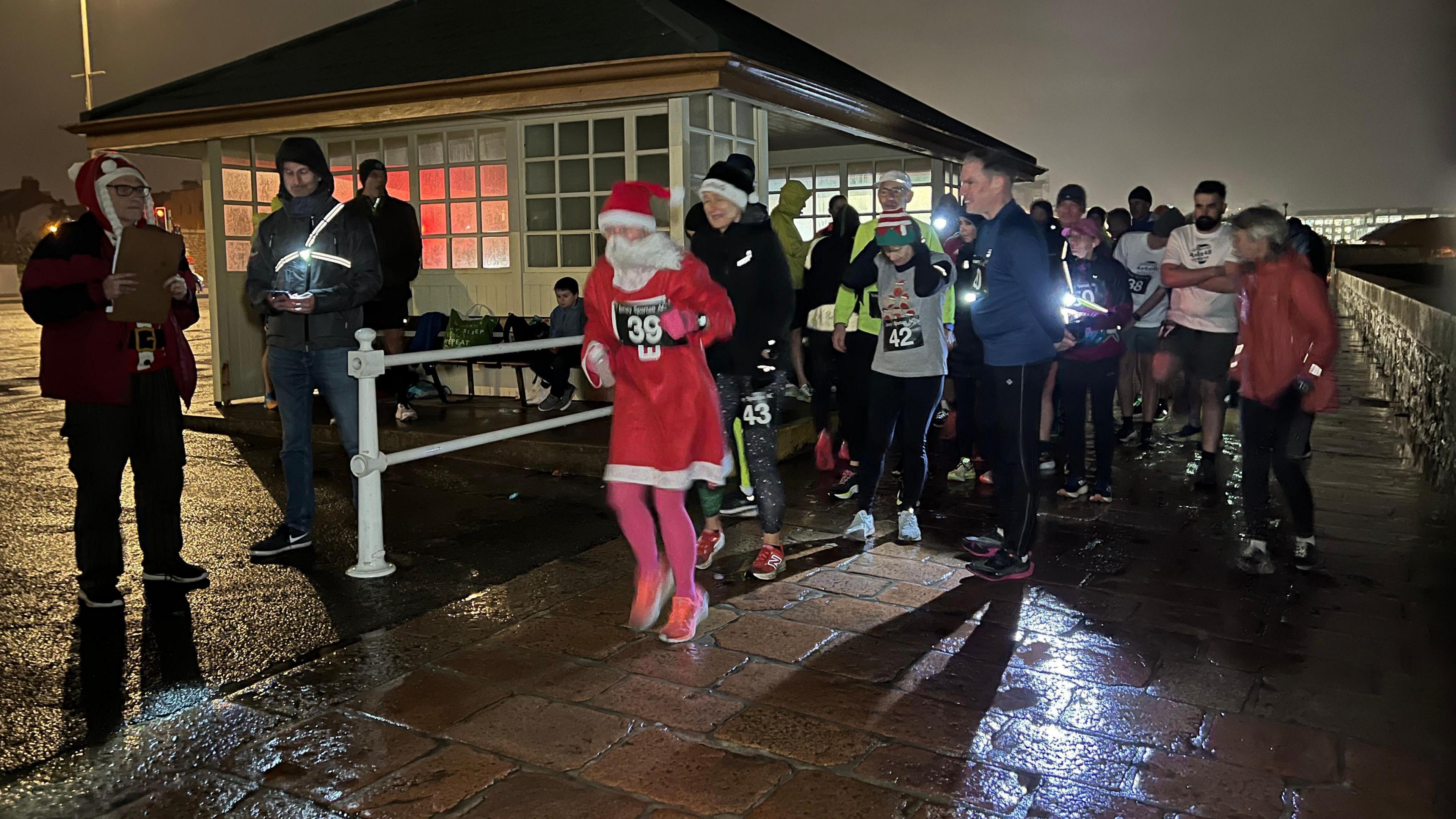 Runners are lined up on a cold and wet night and many are wearing torches as they get ready to set off. The runner at the front is dressed as Ms Claus and others are wearing festive hats. They're on a walkway by a bus shelter and a man is holding a clipboard while another man is using his phone to time when the runners should set off.