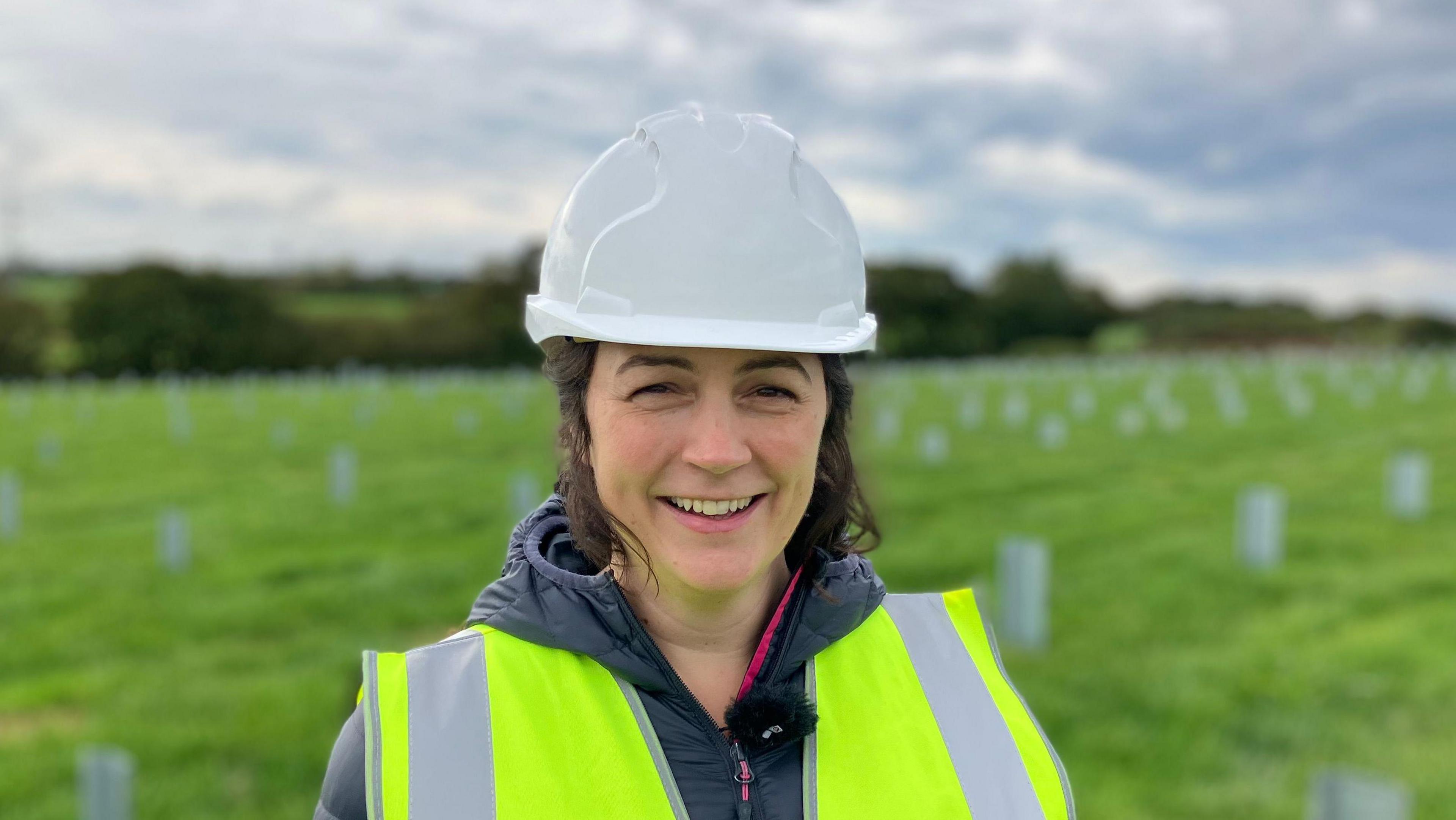 A woman stands in a field in the countryside with construction work in the background. She wears a hard hat and a high-vis vest. 