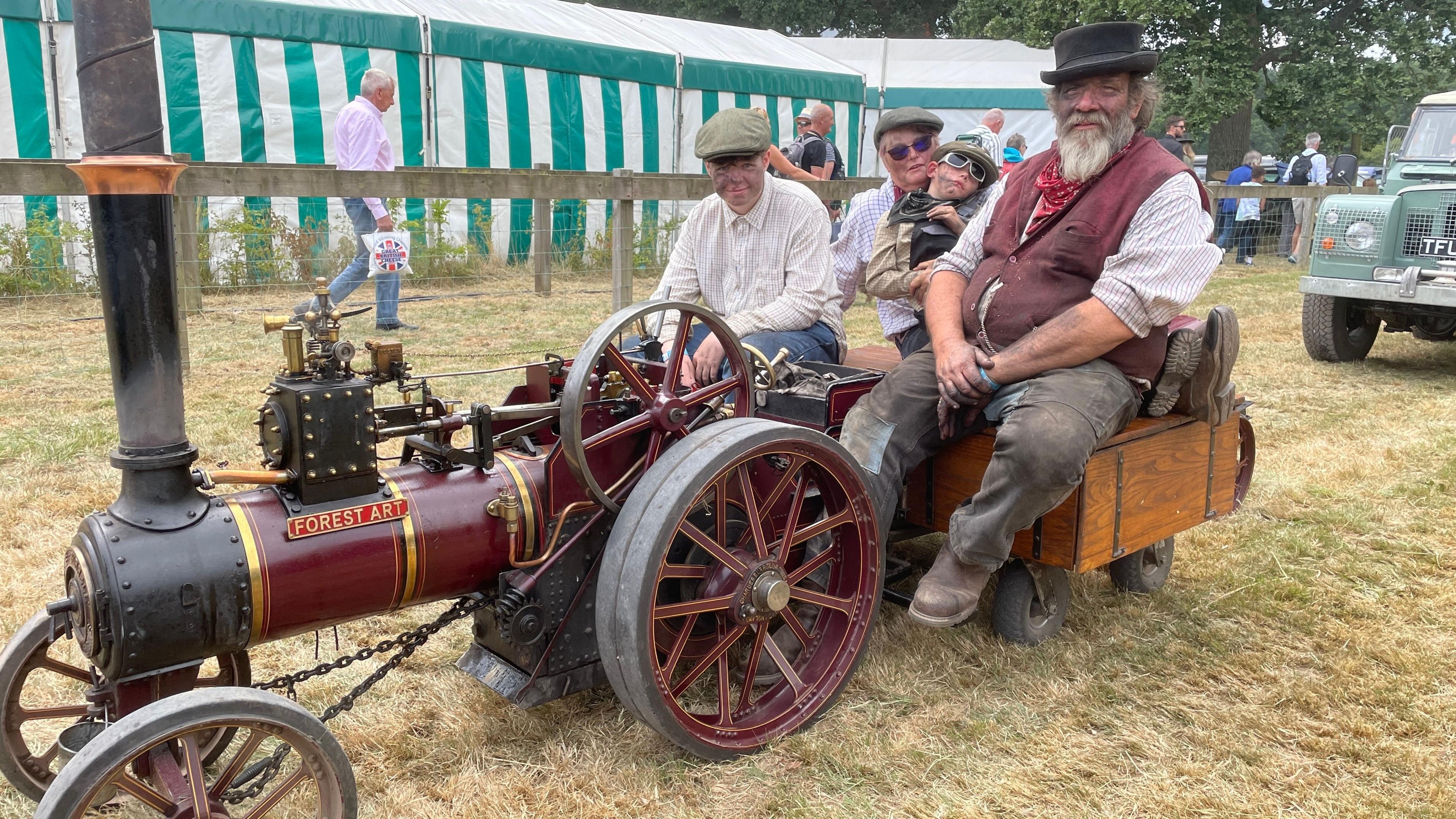 A red retro tractor. Two men, a woman and a child sit on the tractor all wearing flat hats. 