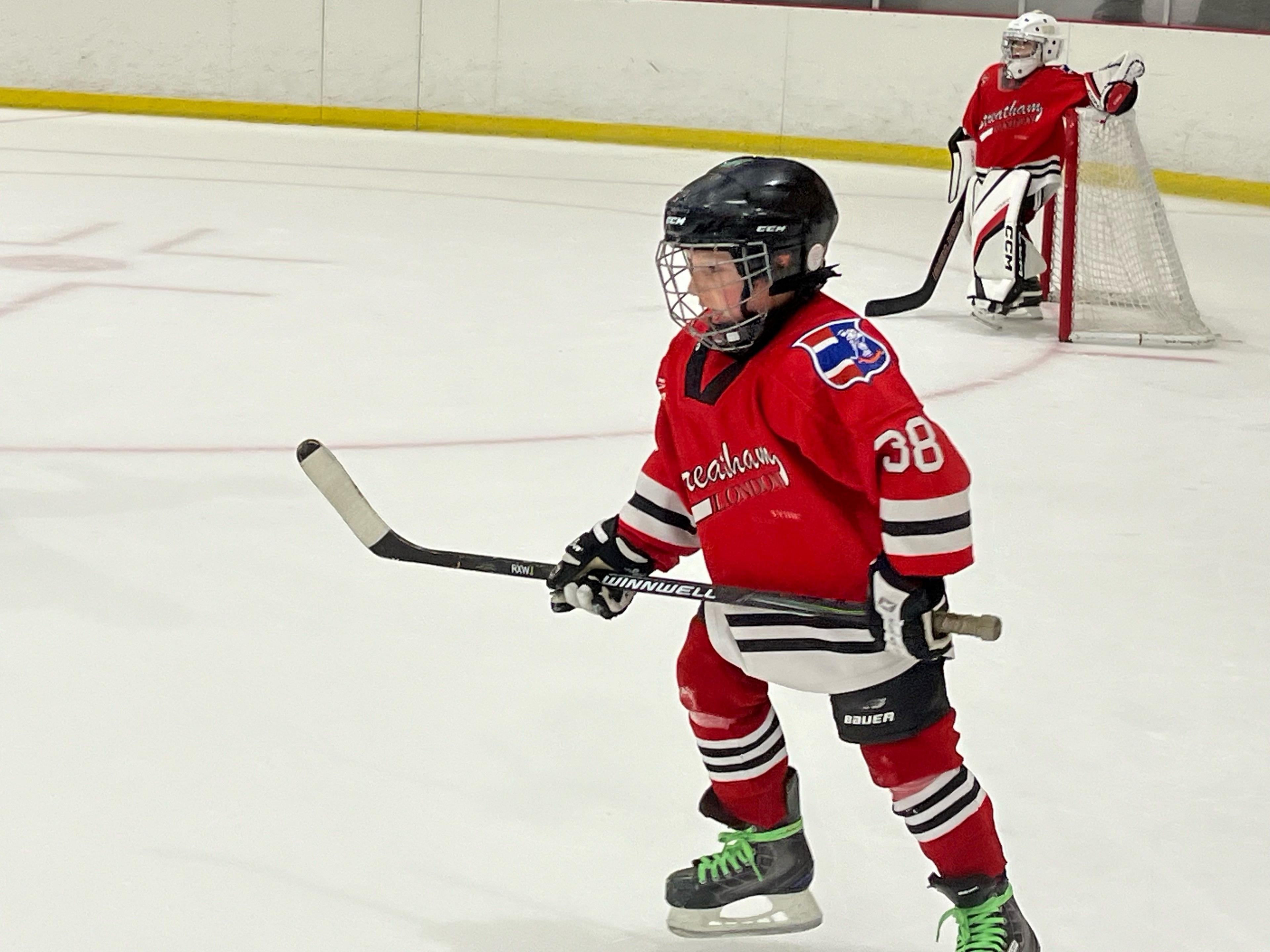 Rufus Quinn, wearing a red Streatham ice hockey jersey with the number 38, skates on the ice during a game, holding his hockey stick. In the background, the team's goalie stands in front of the net.