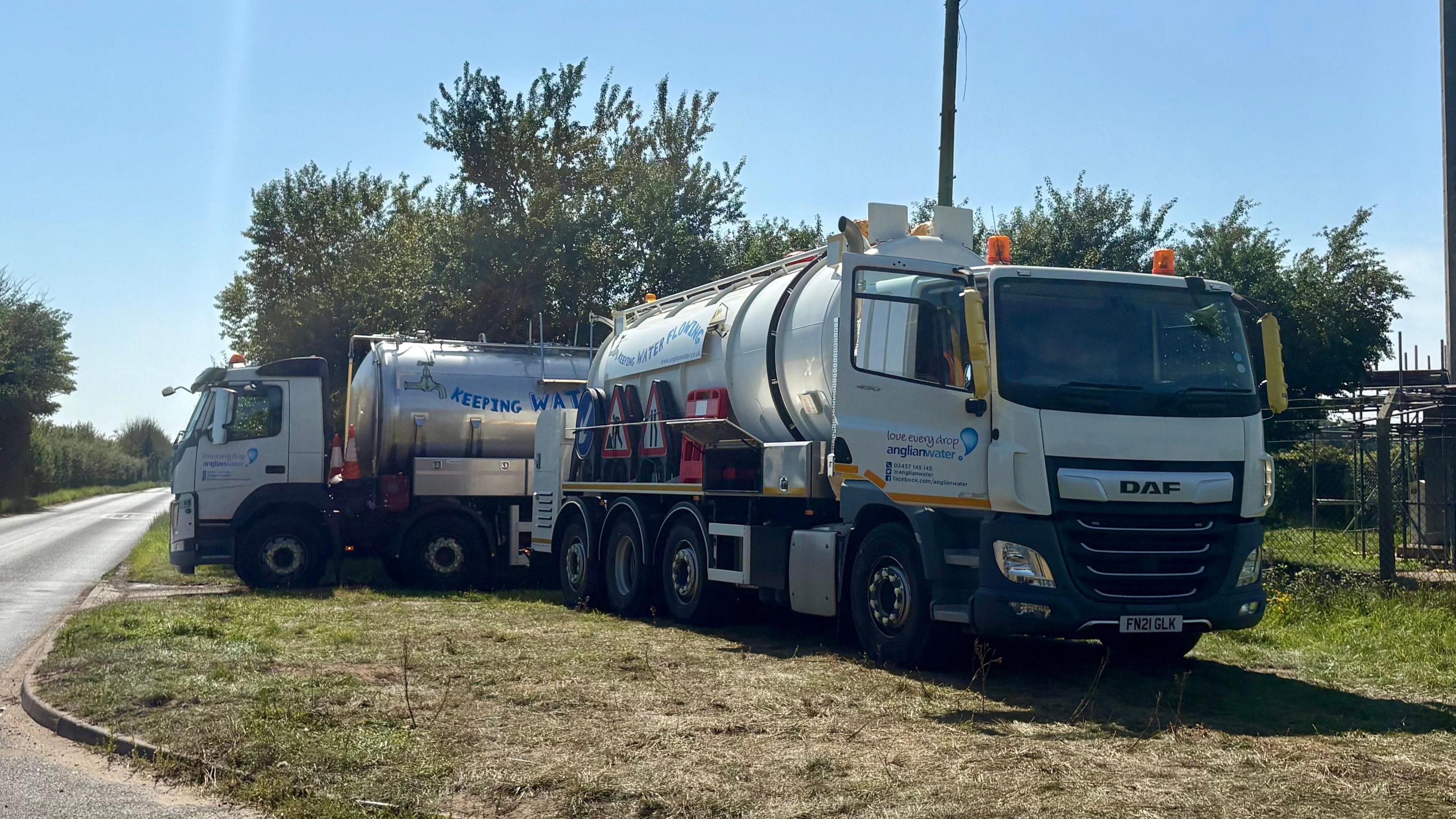 Two Anglian Water tankers are parked on grass just off the side of the road