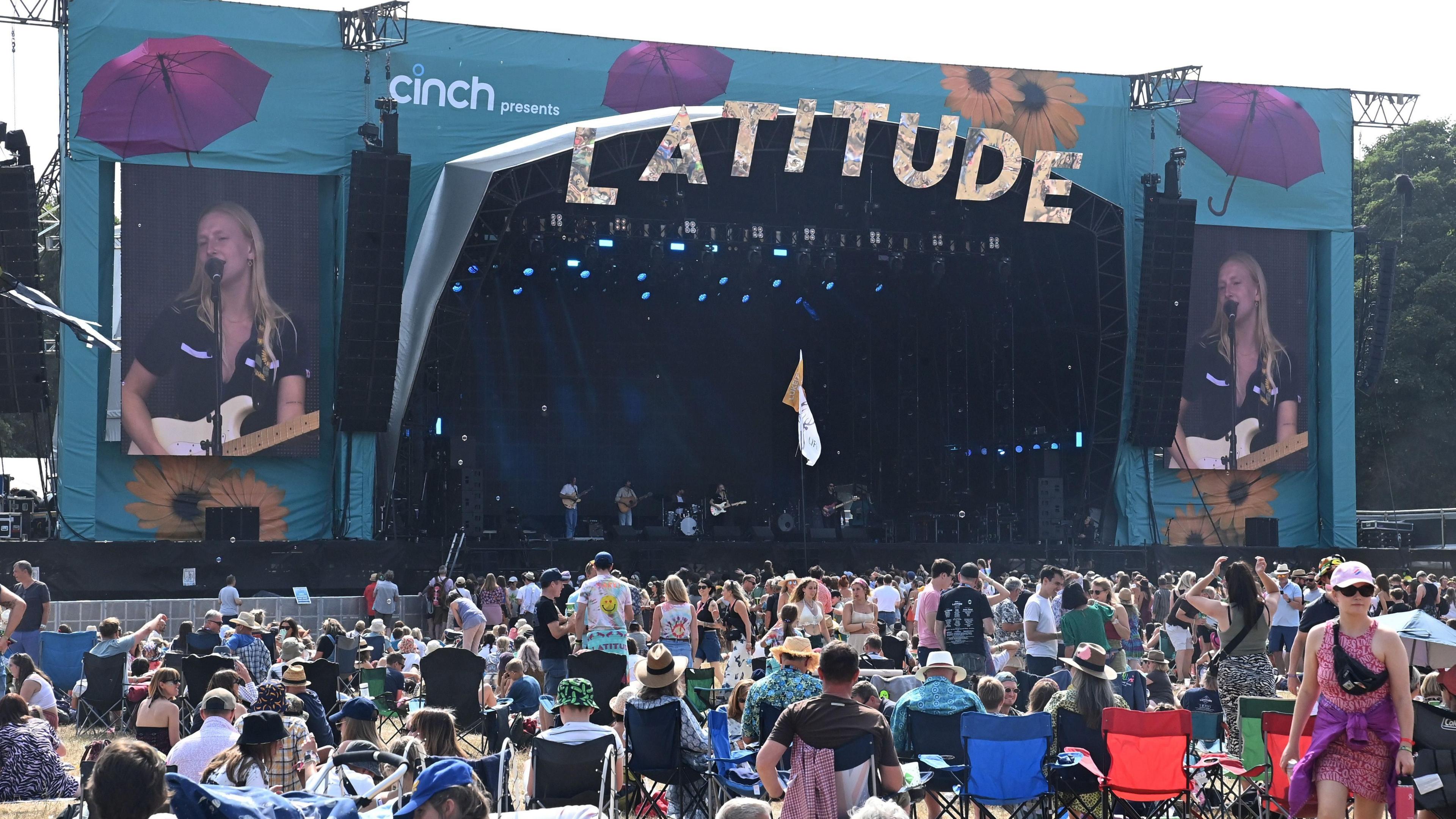 Billie Marten, who has blonde hair and is wearing a black top, performs on a large blue stage during Latitude 2022. There is a busy crowd watching on.