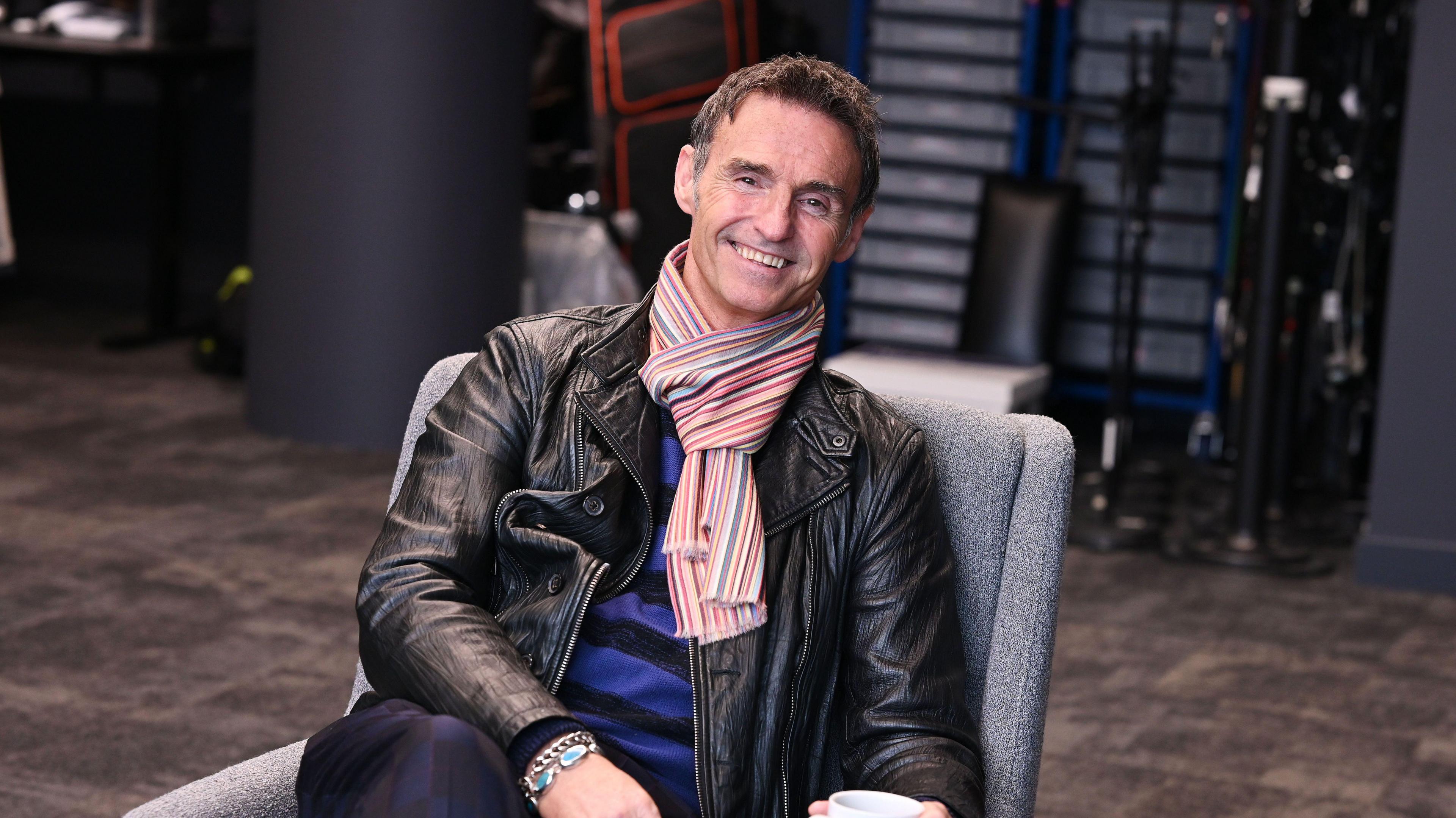 Marti is sitting on a grey lounge chair and smiling at the camera. He is wearing a black leather jacket, blue and black top and a stripy scarf. He has short brown hair.