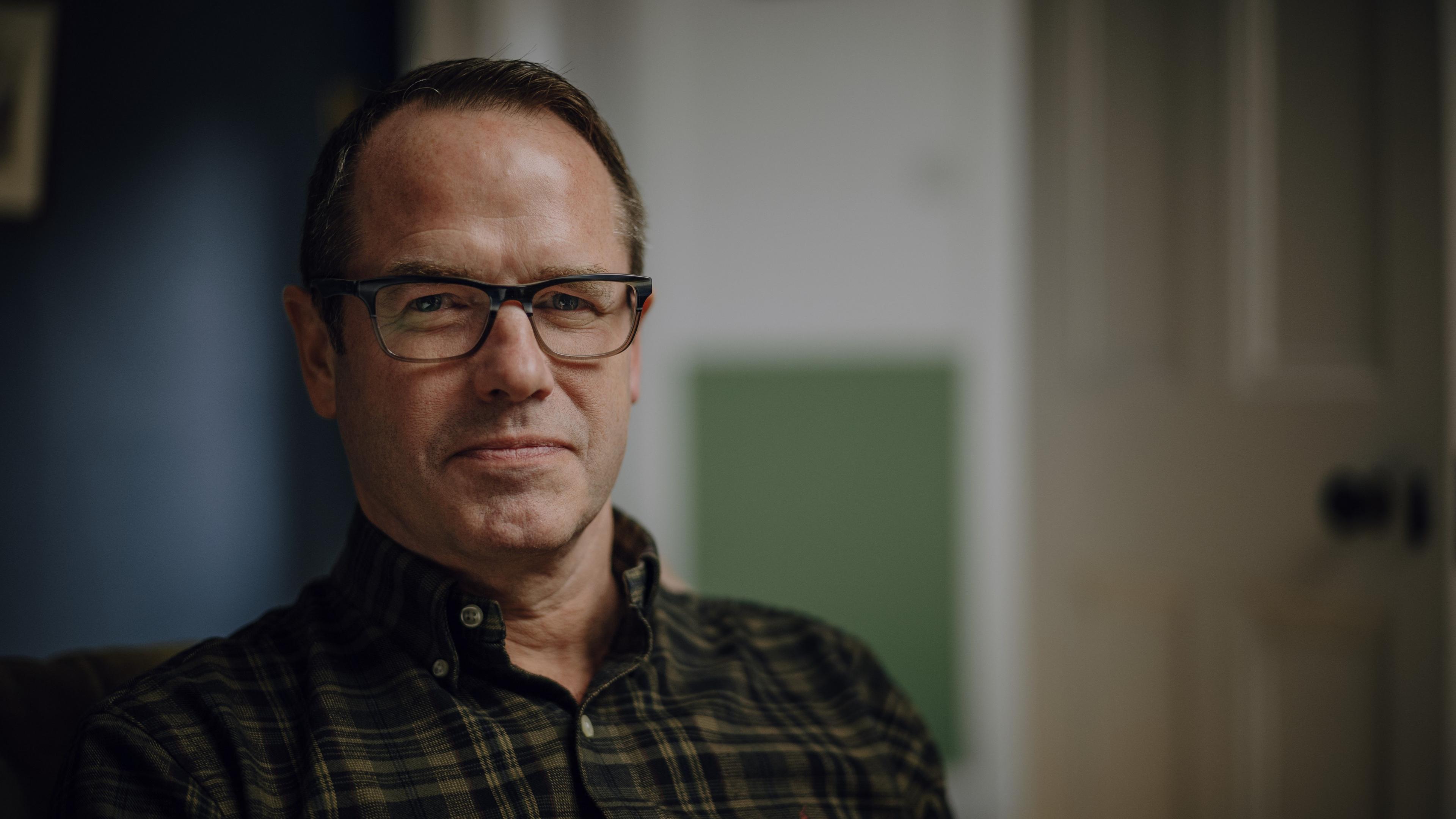 Shane Gorman in a dark checked shirt and dark-rimmed glasses, is smiling to camera in a blue and green room. A wood door can be made out, out of focus, in the background.