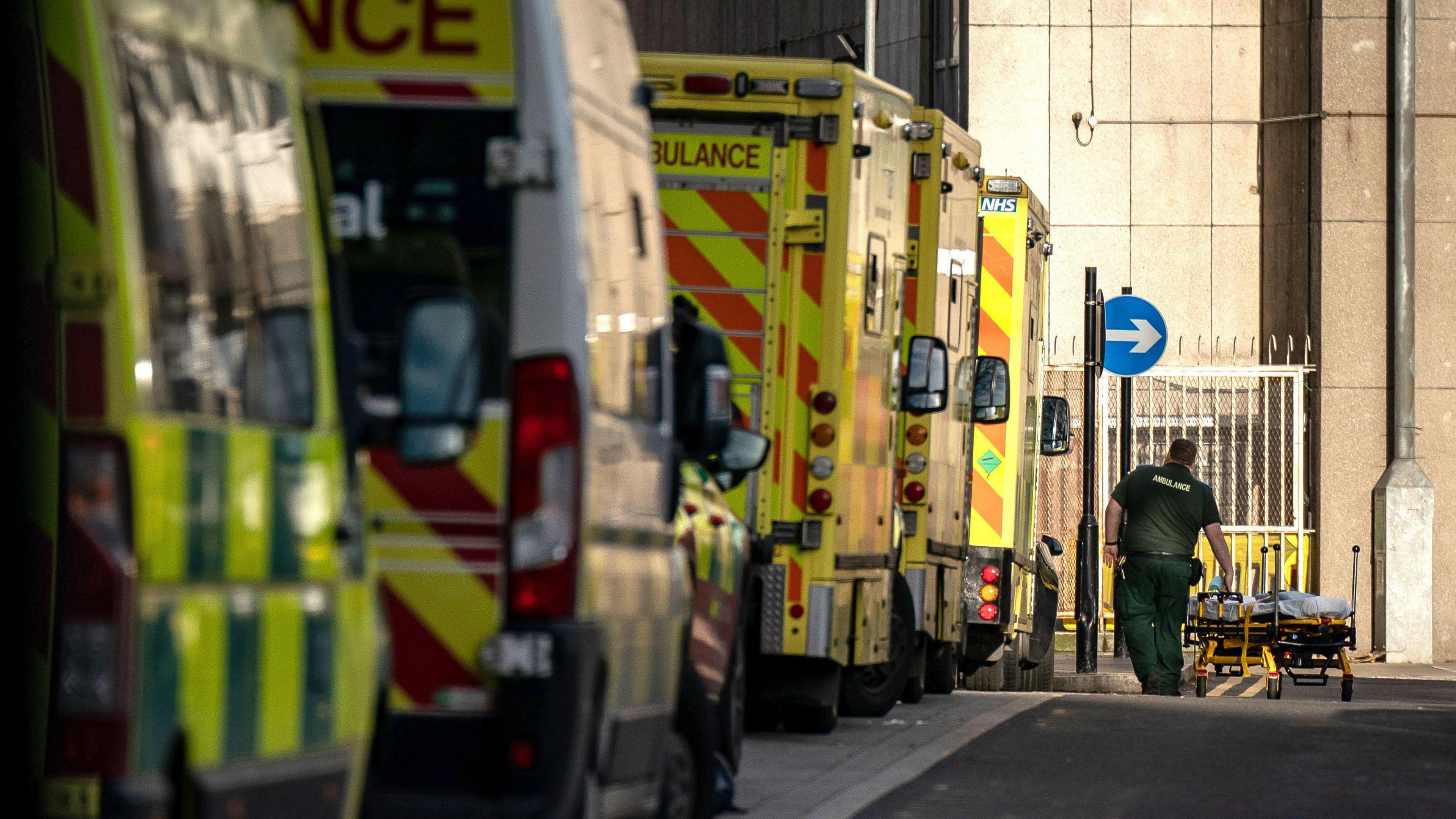 A row of ambulances queuing to get into a hospital. A paramedic in the distance is pushing an empty stretcher.