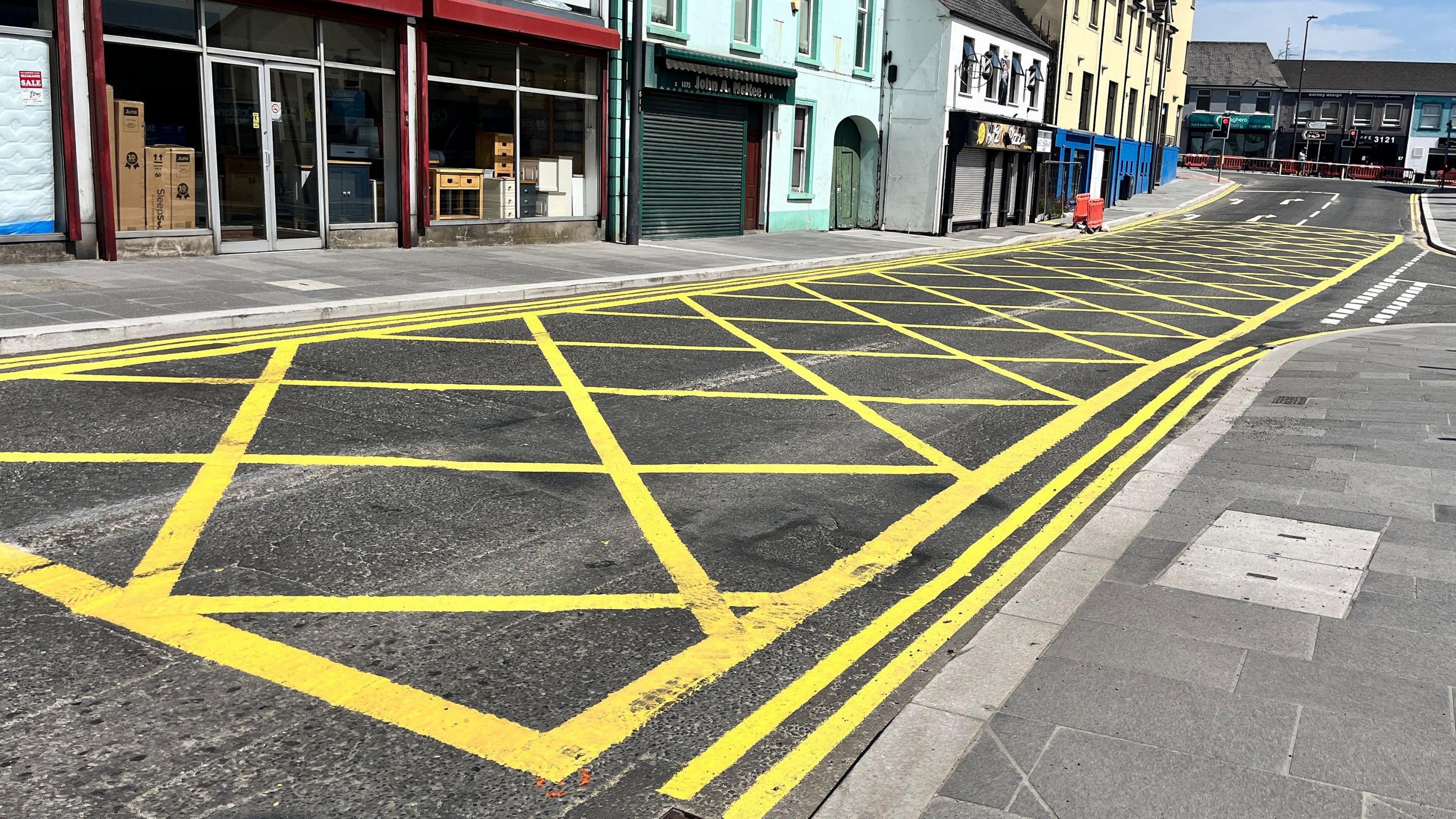 A large yellow box junction leading to traffic lights at a T-junction. Shops line the side of the street.