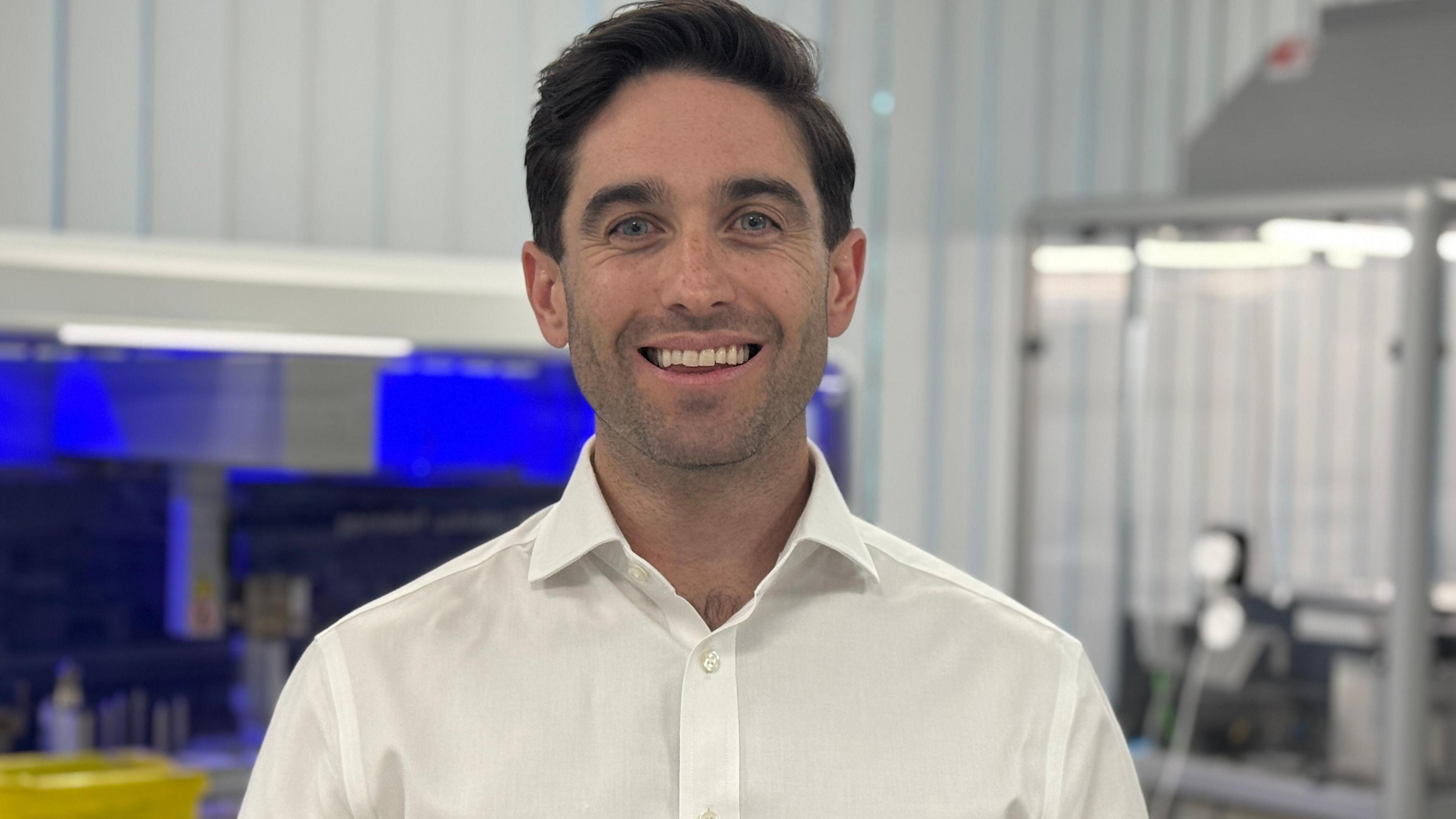 Jeff Bousfield is wearing a white shirt. He is standing in the lab smiling into camera.