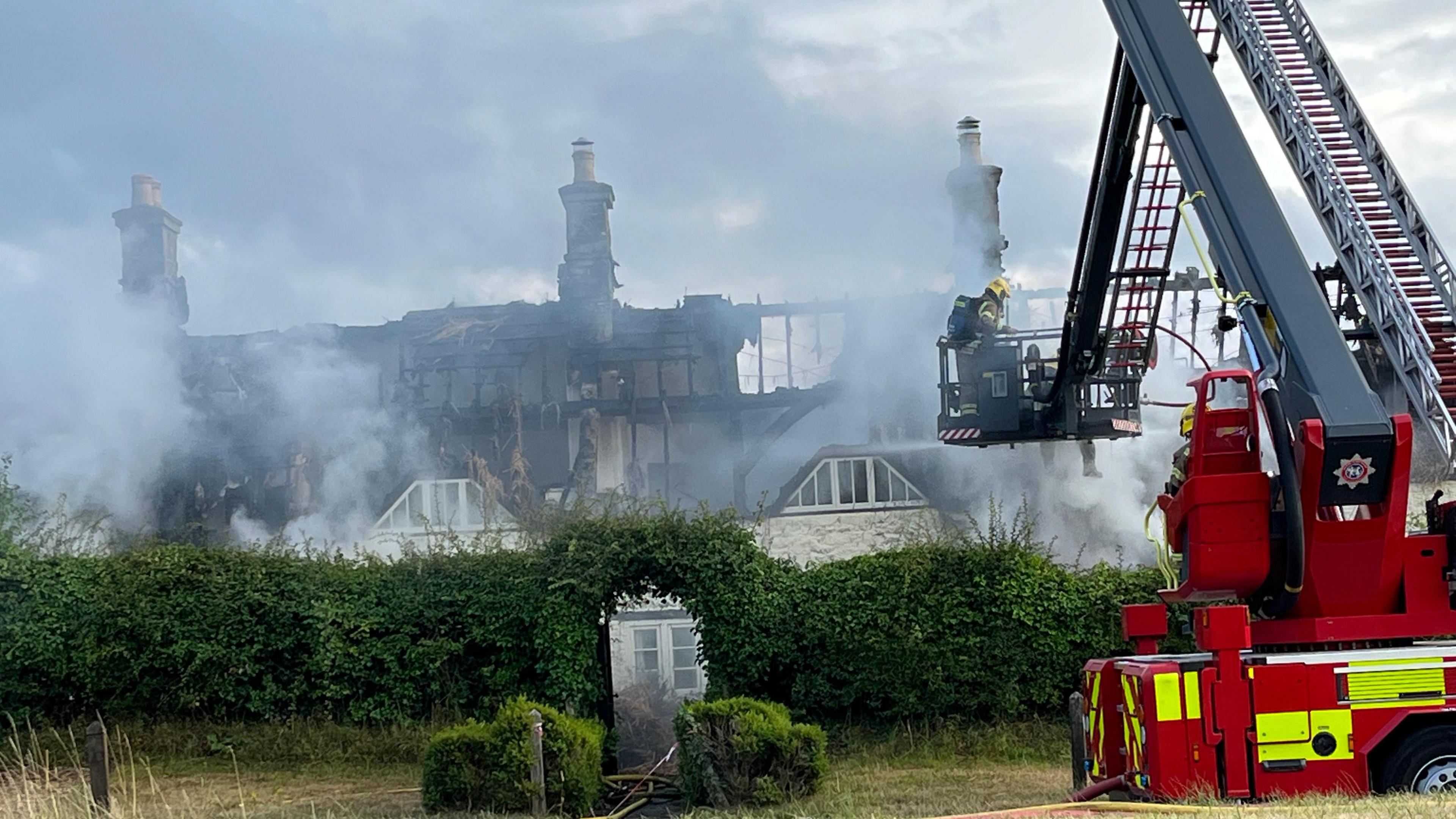 A smoky atmosphere as a fire engine with specialist lift holds firefighters up to the cottage to spray it with water. The top floor is destroyd, with chimneys and the charred skeleton of the timber frames for the thatched roof left