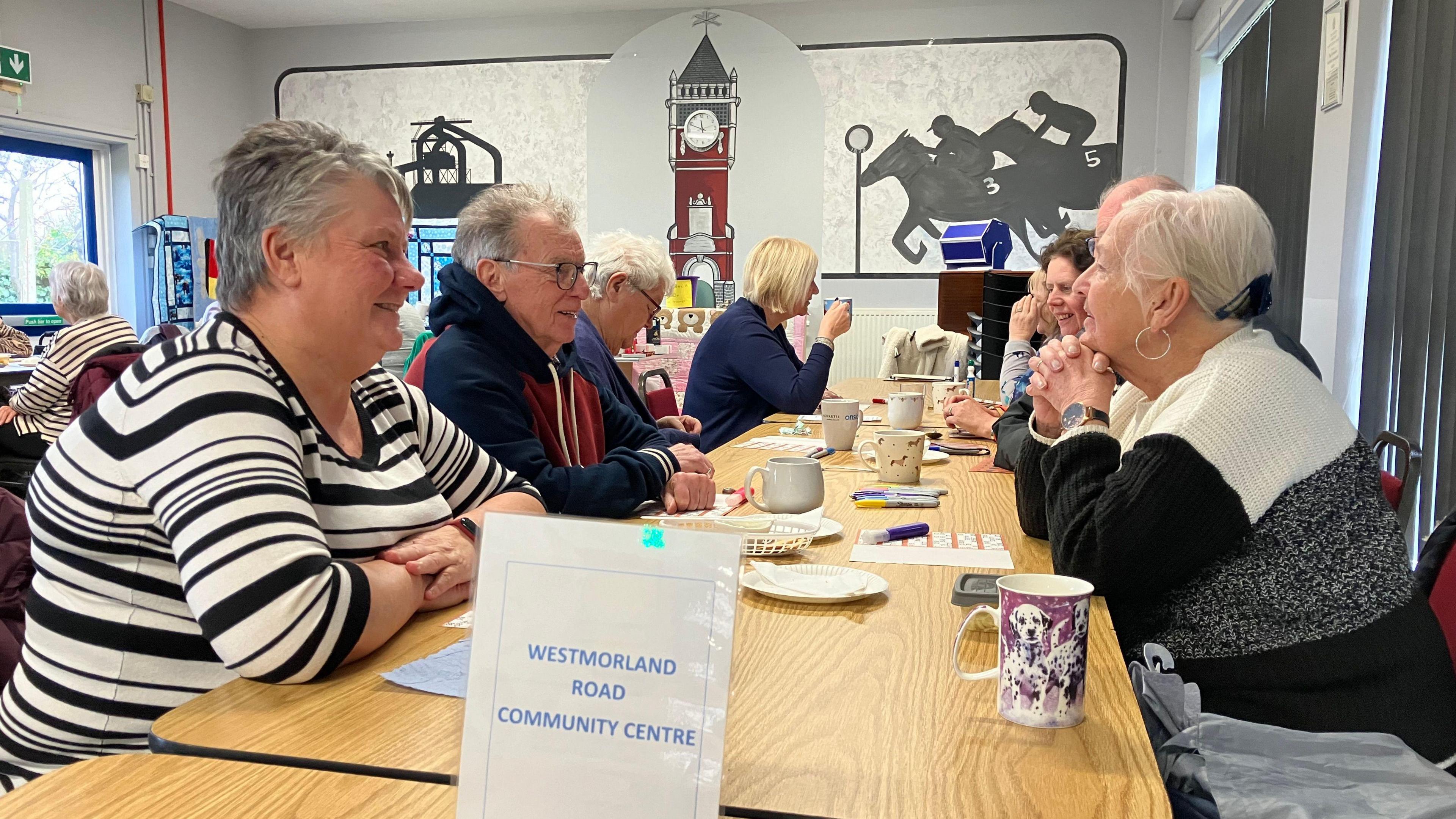 Residents chatting and having something to eat and drink while sitting at a long table.