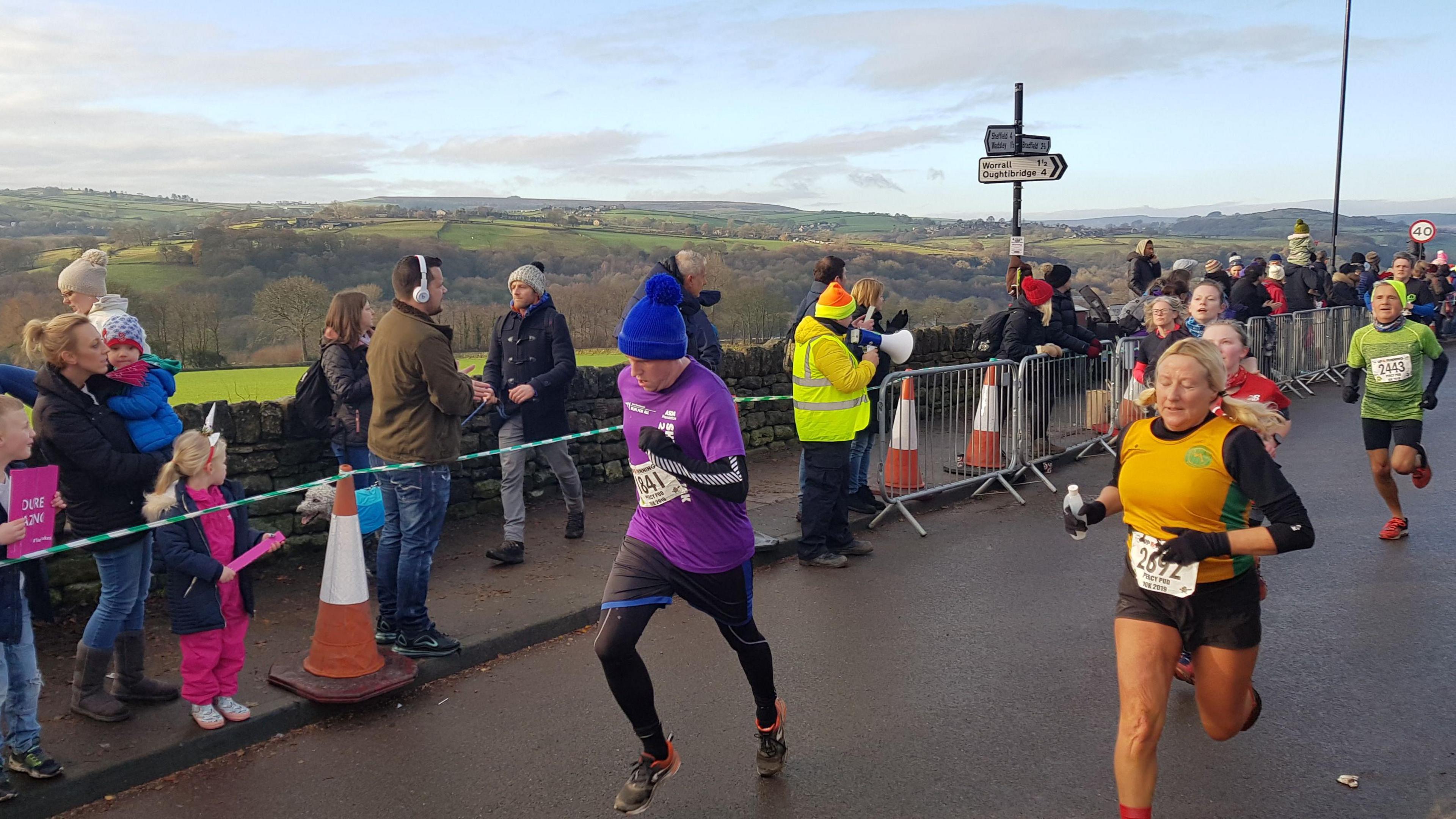 Participants are running down the road, with spectators cheering behind metal barriers on the pavement. In the background are green, rolling hills. In the centre of the photo, one runner is wearing a blue bobble hat.