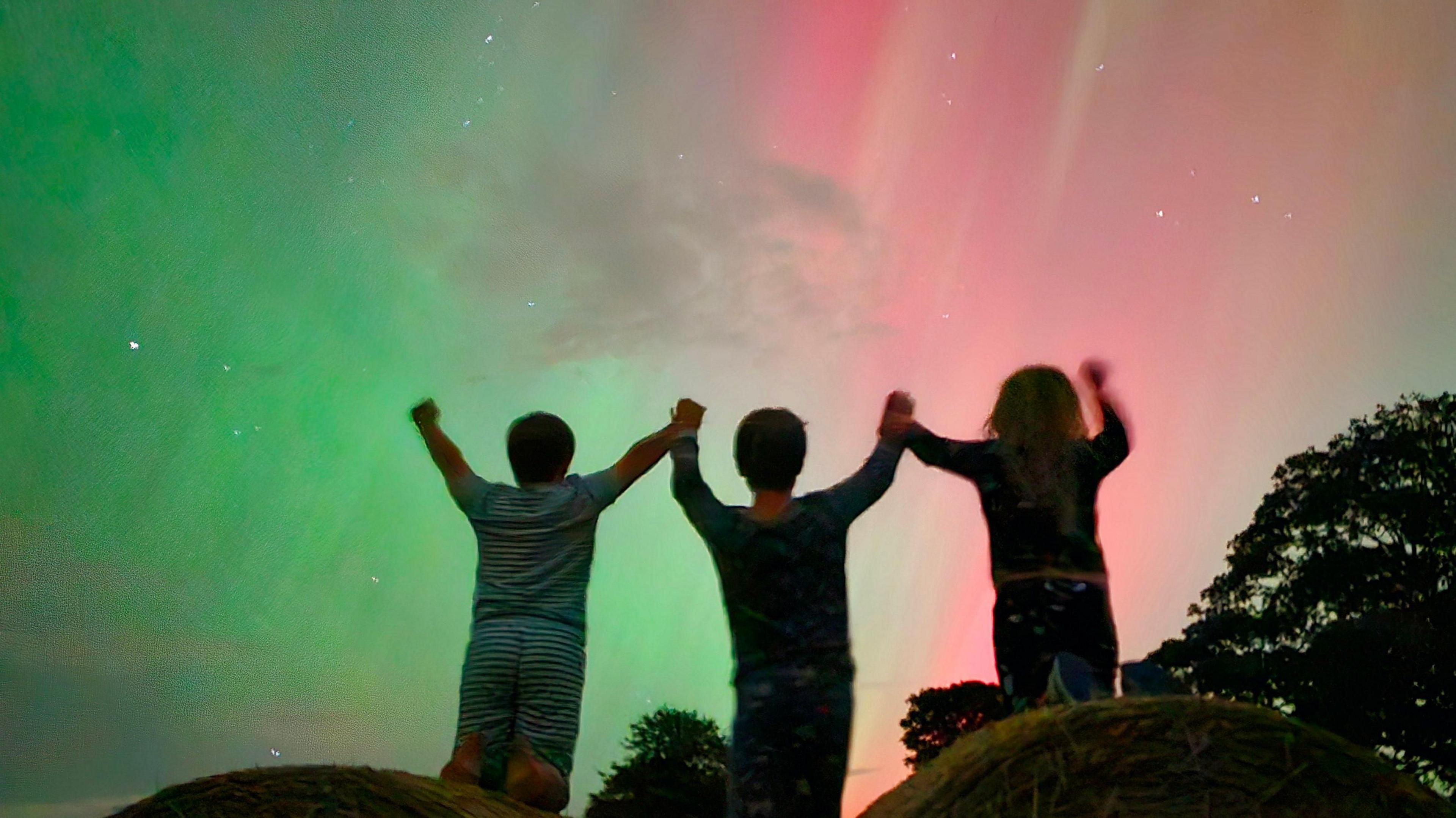 Shadowed outlined of three children, holding hands with their arms raised and the Northern Lights in the sky, showing green and pink colours