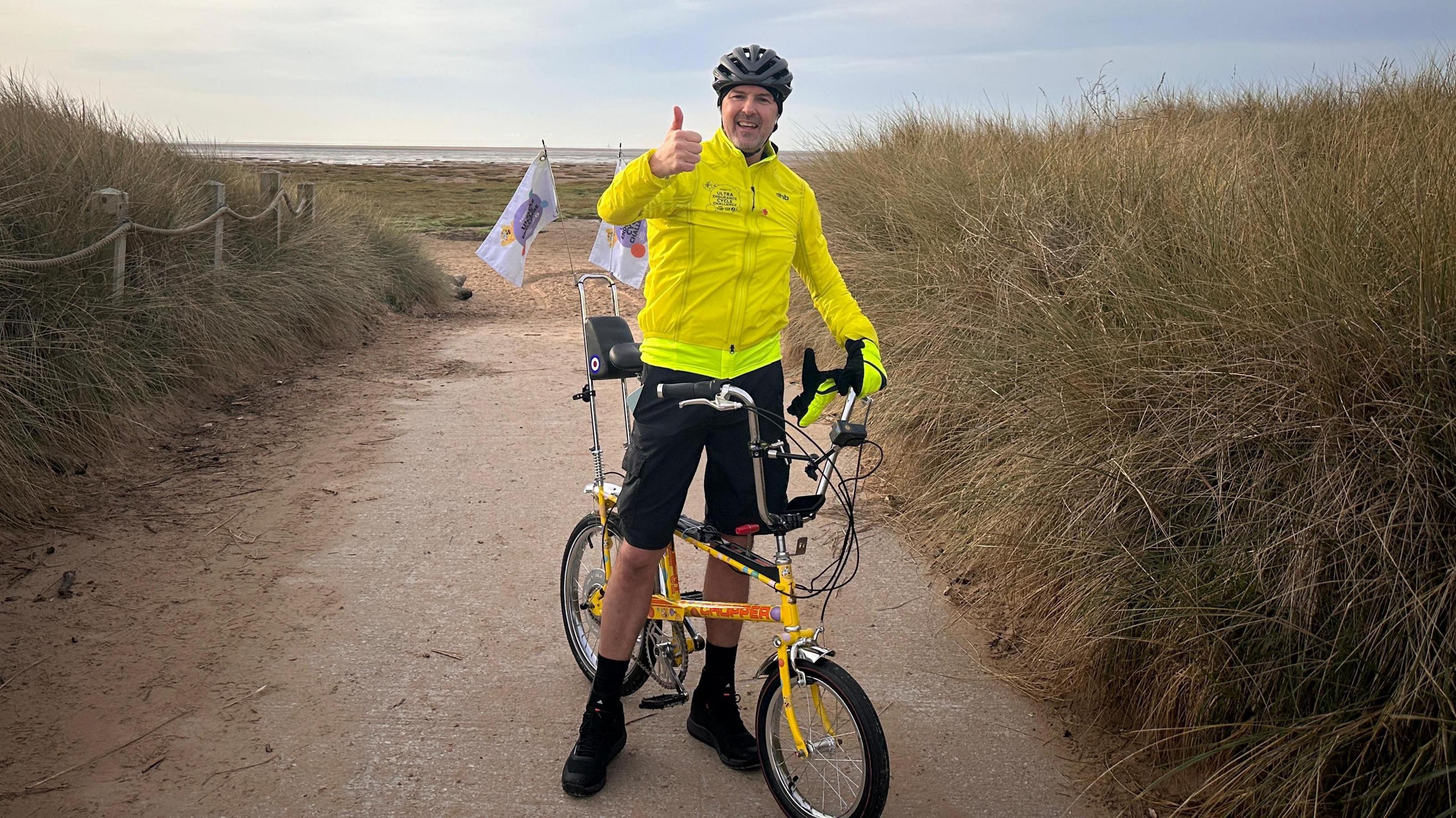 A man wearing a yellow cycling jacket, black shorts and a black helmet standing with a yellow bike giving a thumbs up. The background is a beach and the sea, on either side of the man is long grass.