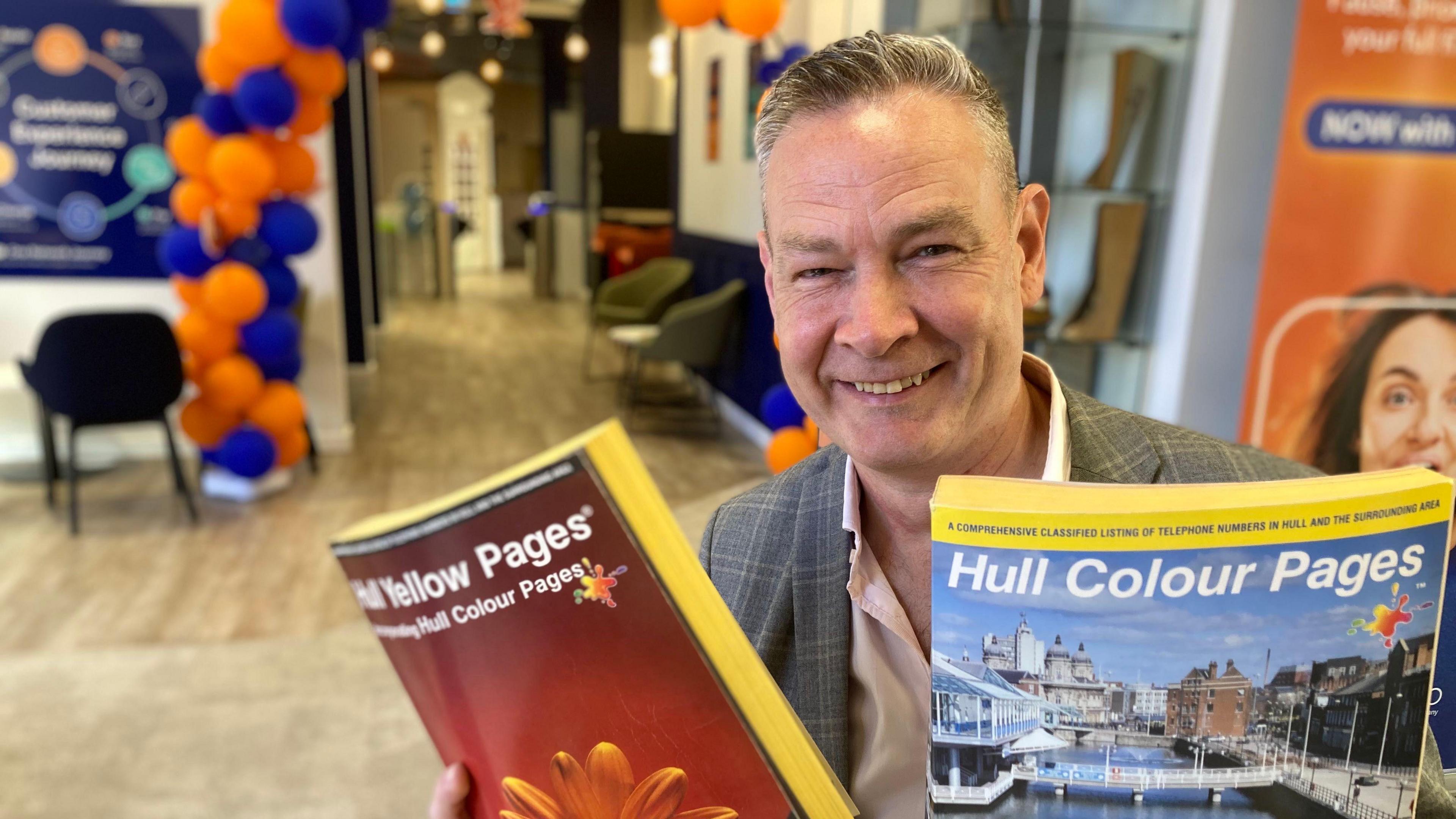 Neil Bartholomew holding a copy of a Hull Yellow pages in his right hand and a Hull Colour pages in his left hand - they are A4 sized books with yellow pages and colourful front covers. Neil is wearing a checked jacket and pink shirt, he has close cropped silver hair and is smiling.