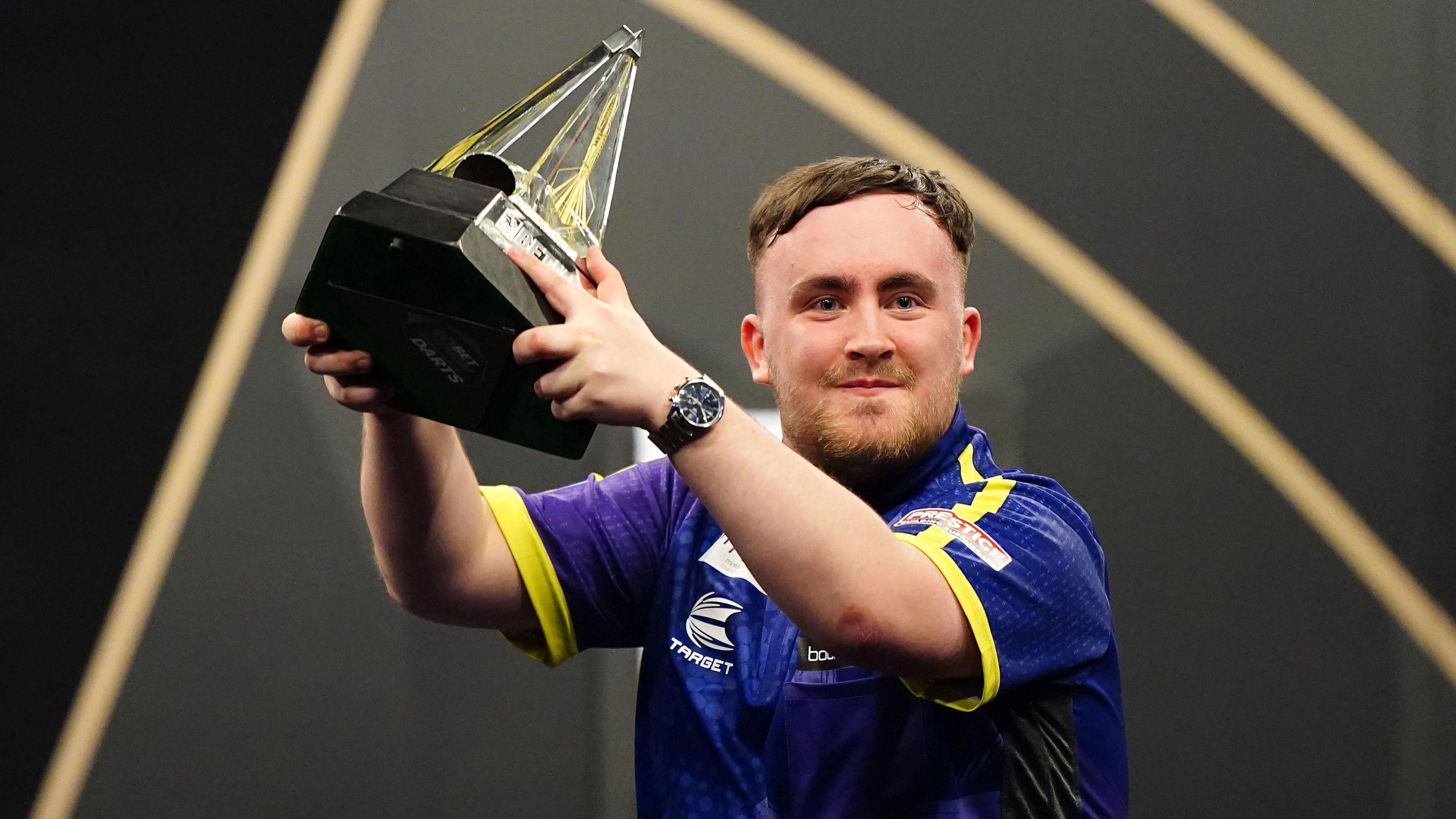 Luke Littler holds up a trophy and smiles