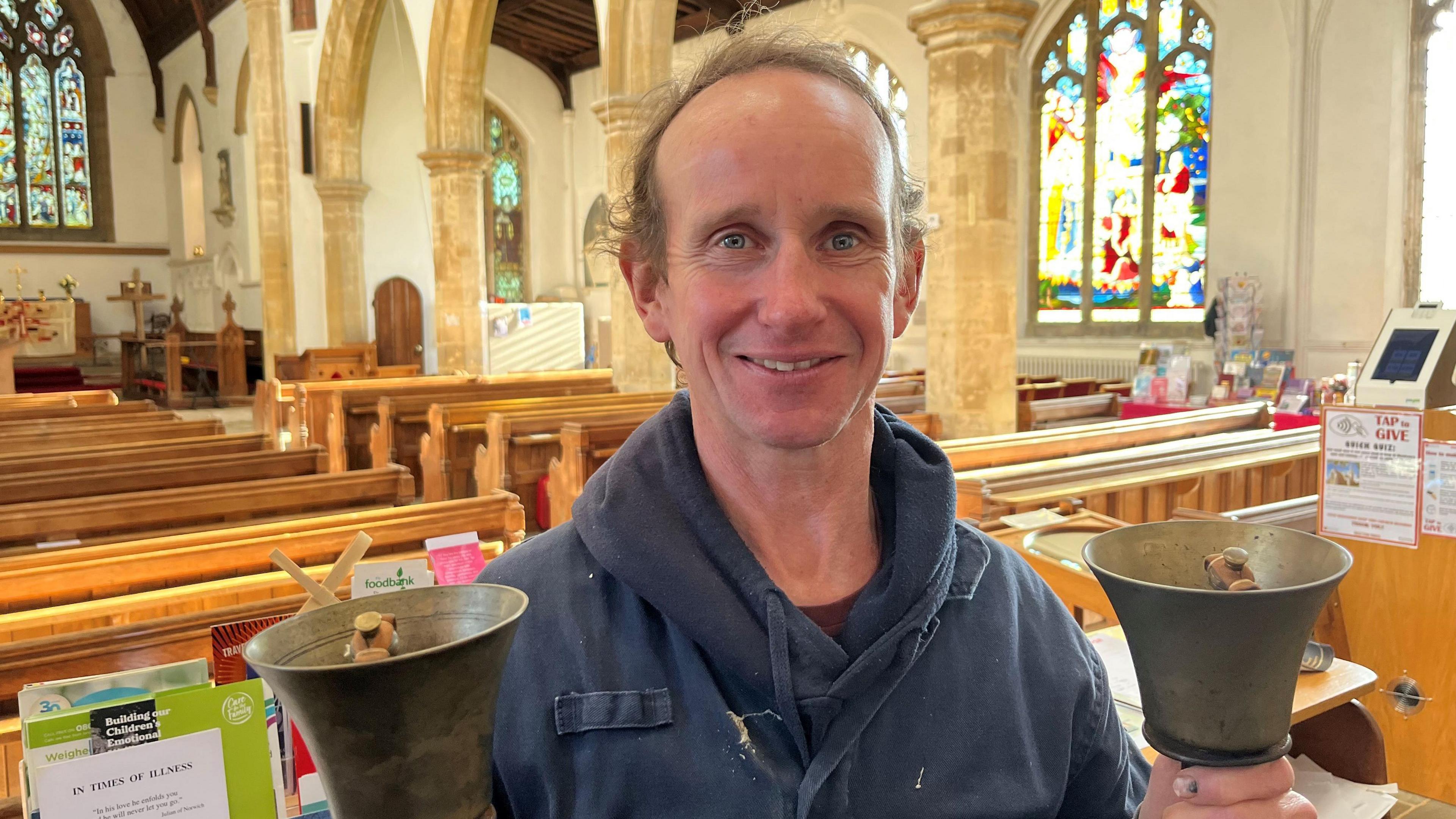 Andrew Goakes smiling at the camera while holding two hand-bells inside the church. He has thinning brown hair and is wearing a grey fleece. Behind him are the pews of the church and a colourful stained-glass window is visible over his shoulder.