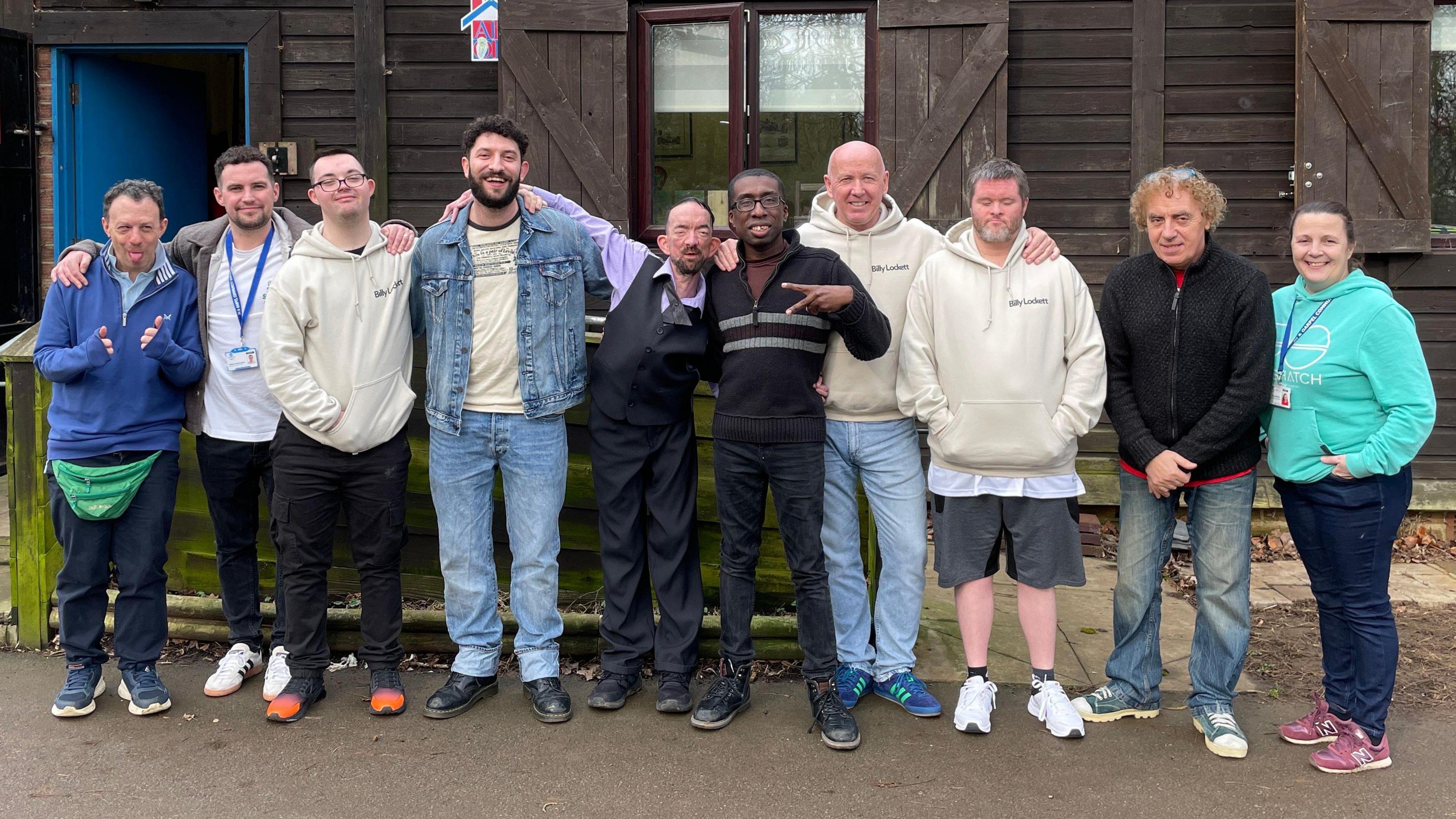 Clients of the JH and Oarpel group are lined up with group leaders outside a brown hut posing for a photo. They are all looking at the camera, casually dressed. In the centre of them is the singer Billy Lockett. Some of them have their arms around each other. 