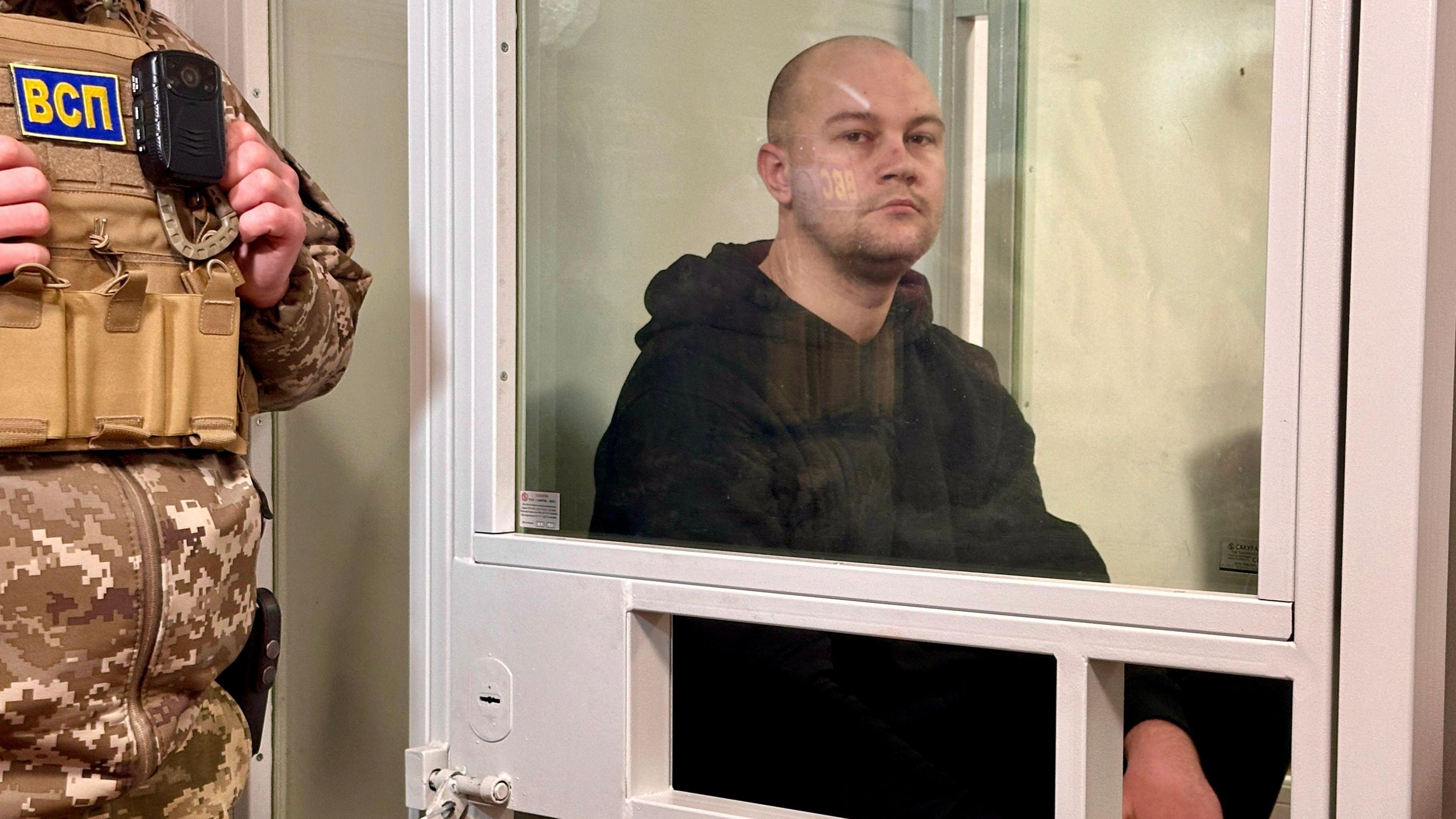 Serhiy sits inside a glass cage wearing a black hoodie, with a uniformed guard standing outside