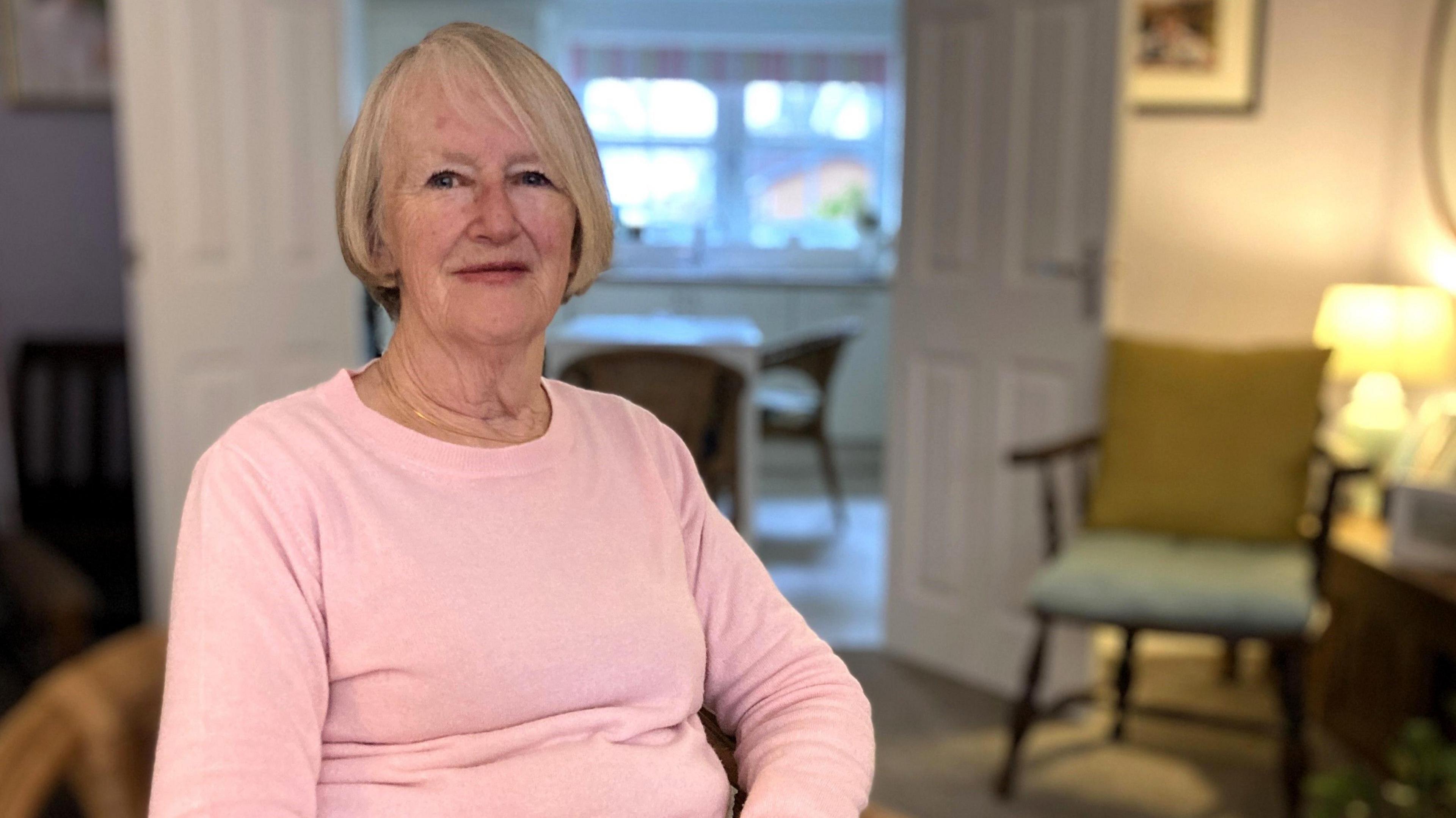 Margaret Parish sat in her living room. A lamp is on in the corner of the room and she is sat in a chair with a pink top on. She has cropped grey hair.