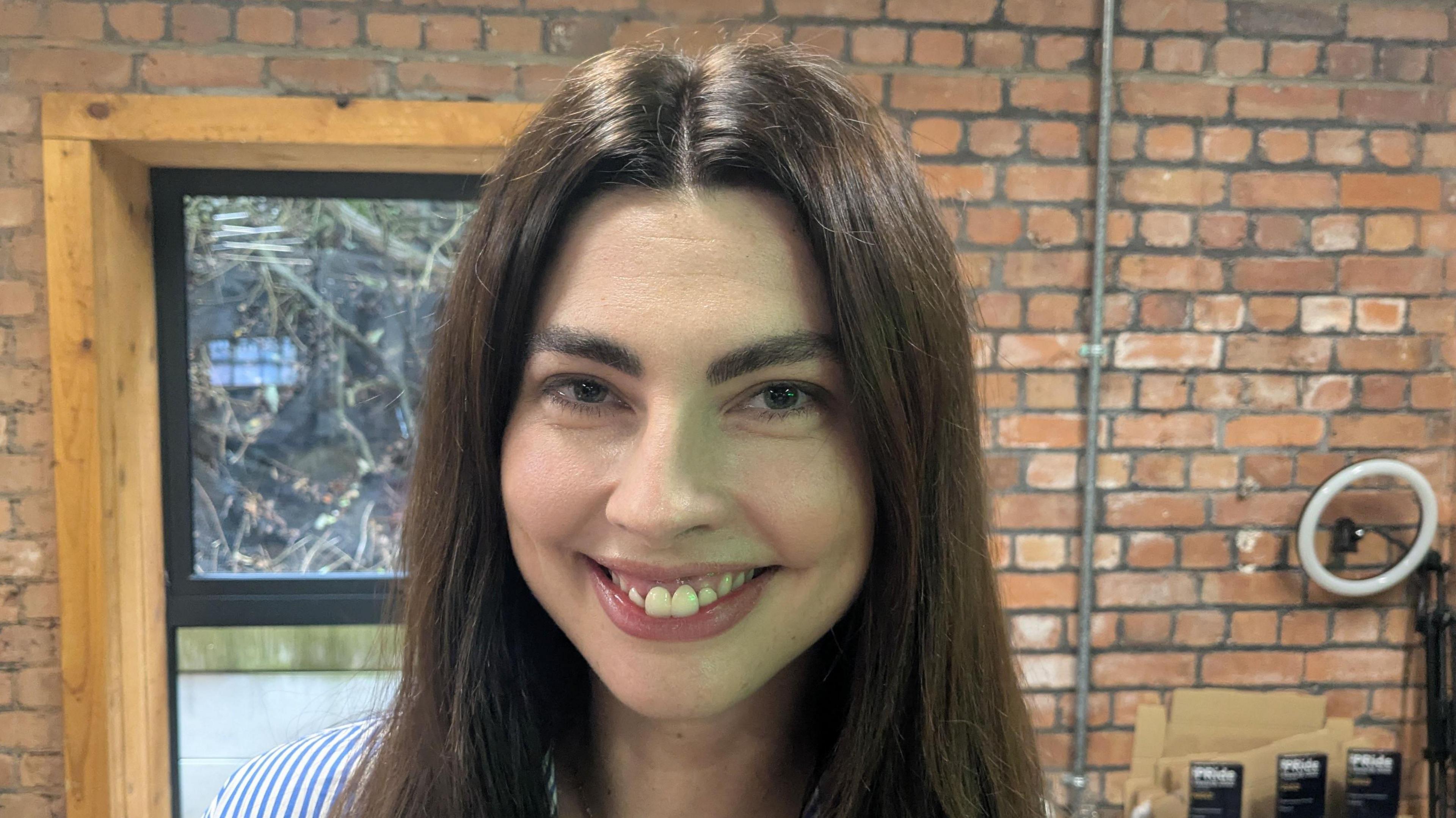 Sophie smiles at the camera. She has longs brown hair and is wearing a blue and white striped shirt. Behind her is a small window and an exposed brick wall. It is a head and shoulders shot.