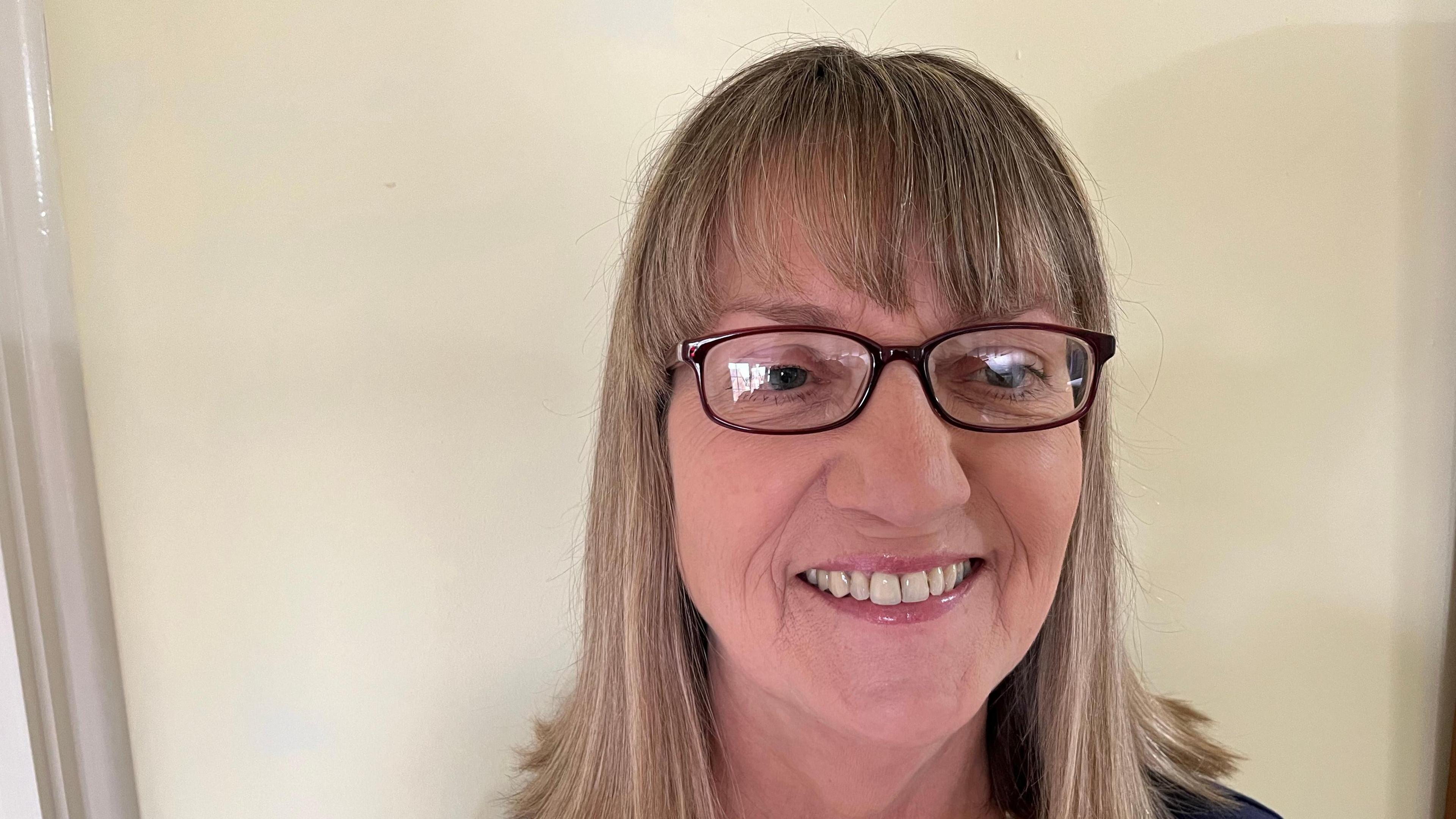 Jill Bolton, 63, has sandy shoulder length hair and has glasses with dark frames. A magnolia coloured wall is behind her.