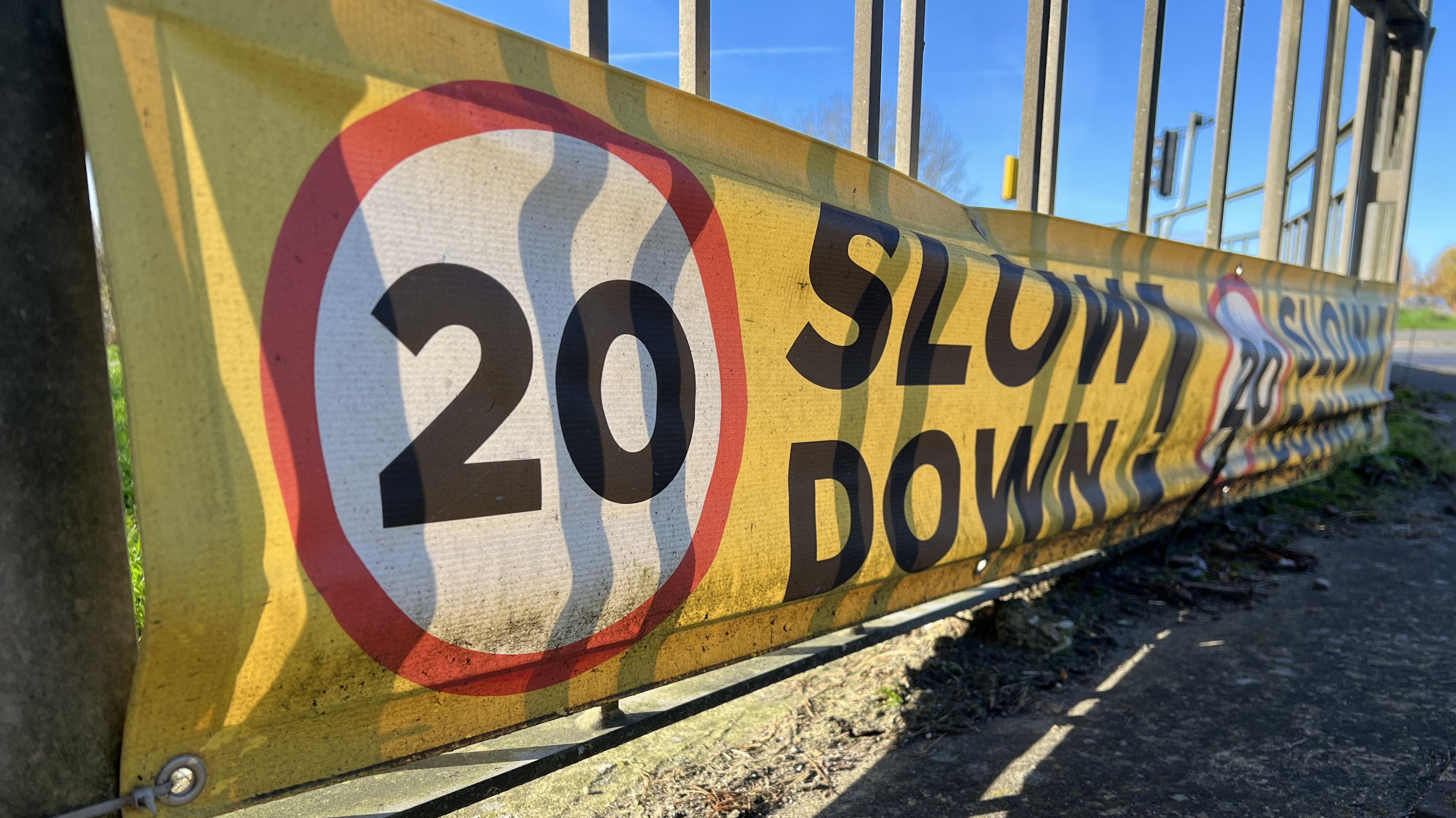 A yellow and red sign that reads "20 slow down". It is attached to railing near Nova Hreod Academy.